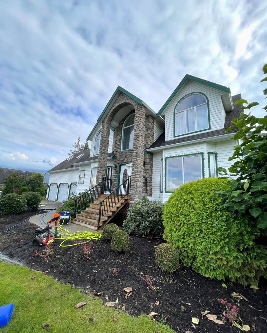 A man in yellow rubber boots operates a pressure washer to clean a patio surrounded by vibrant greenery and colorful flowers under a clear blue sky.
