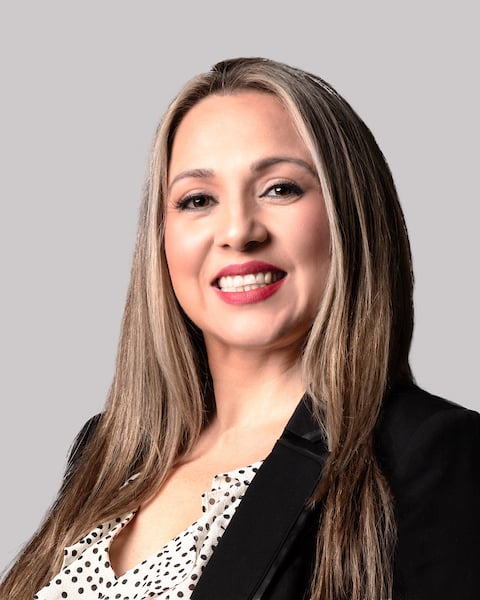 Smiling woman with long blonde hair, wearing a black blazer and polka dot blouse, against a plain gray background.
