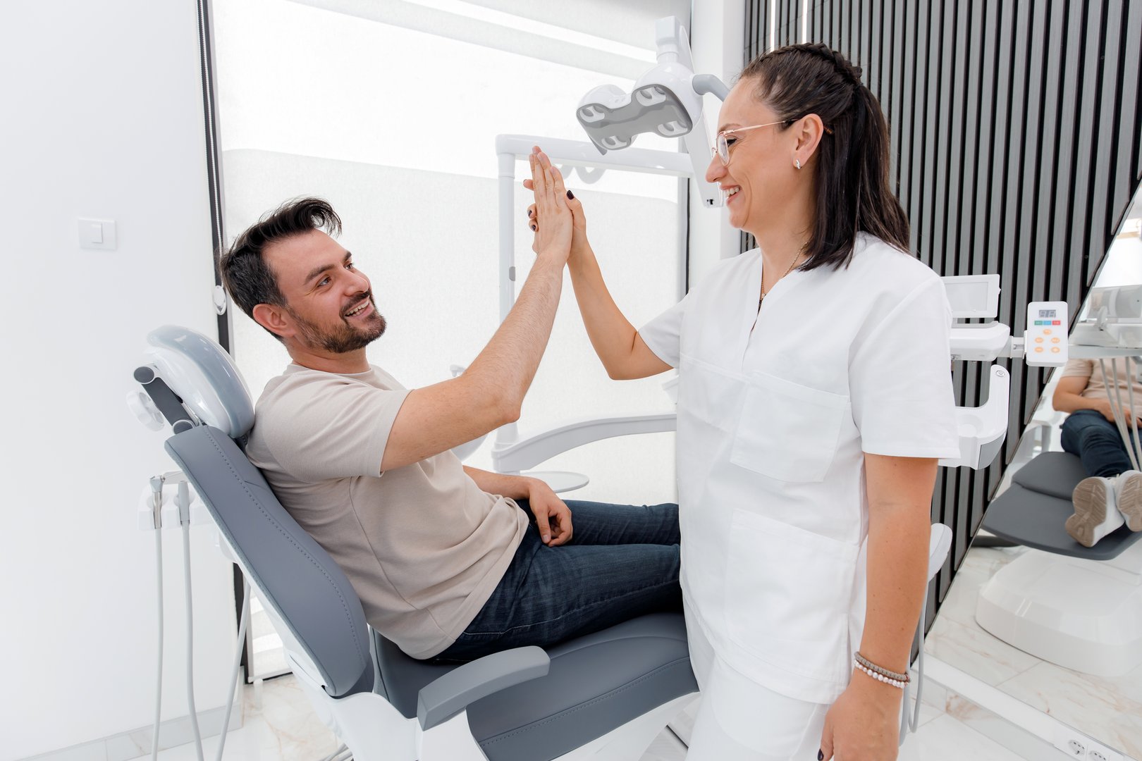 A man in a dental chair happily shares a high-five with his dentist, showcasing a positive atmosphere in a bright, contemporary dental office designed to ease patient anxiety.