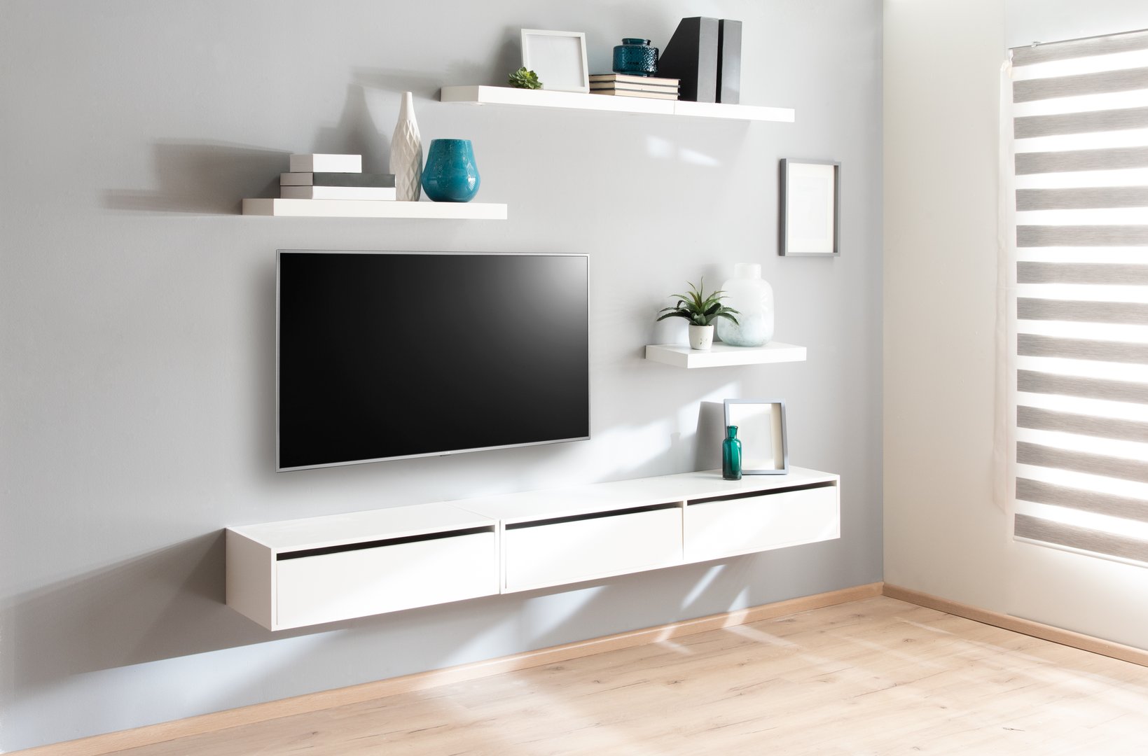 Interior photograph showcasing a sleek, minimalist media wall in a modern home. A large television is centered above a white three-drawer floating cabinet, complemented by white wall shelves holding simple decor against a pale gray wall. Natural light streams in from the window with blinds. Ideal for interior design concepts or furniture marketing.