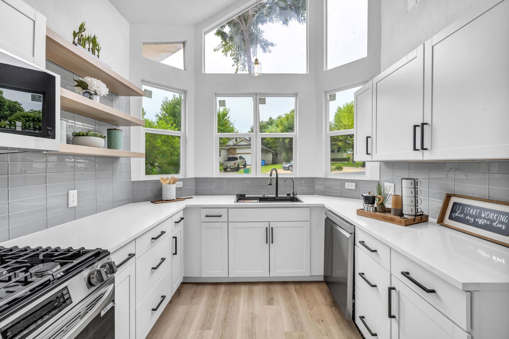 Modern kitchen with white cabinets, large windows, light wood flooring, and a black sink, featuring open shelving and appliances.