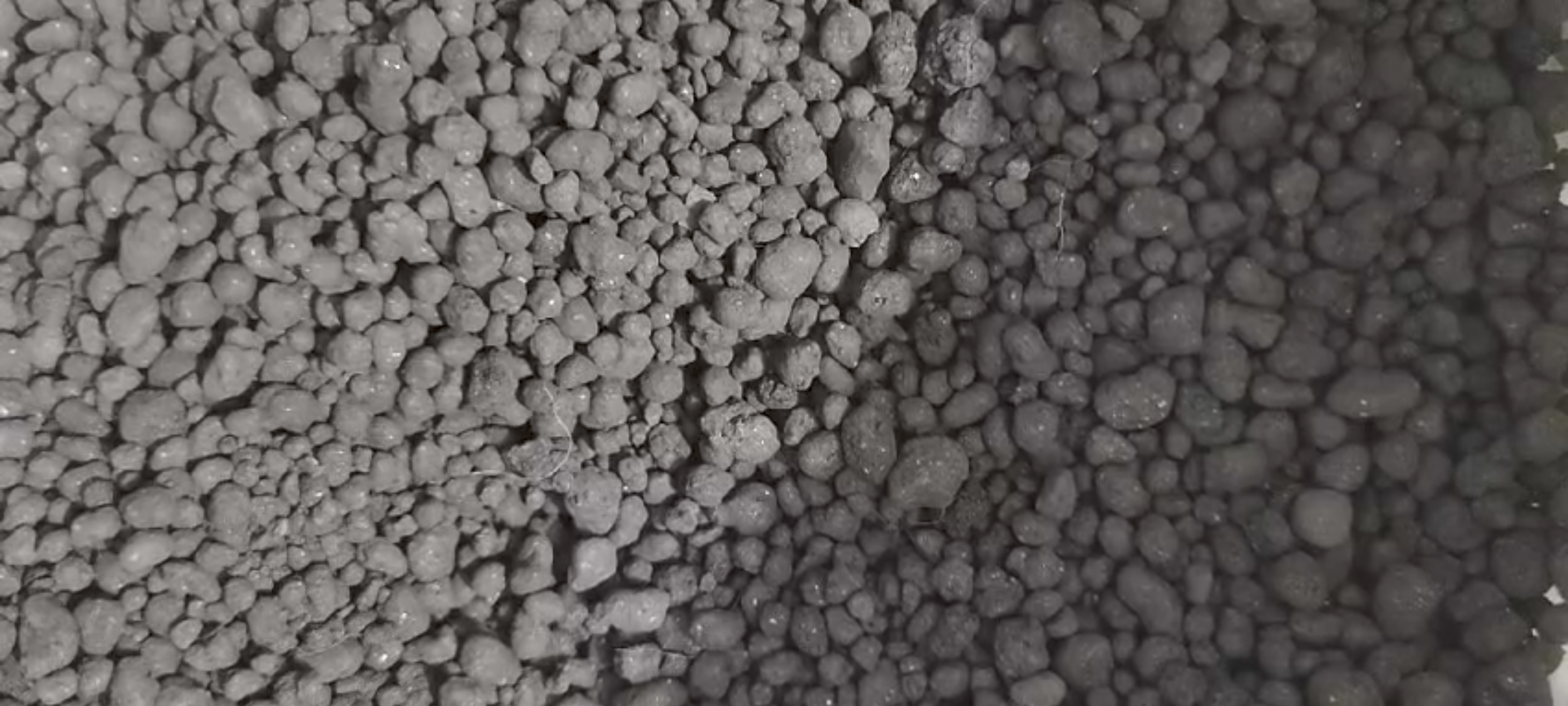 Close-up of a pile of dark gray, round pellets varying in size, resembling fertilizer or industrial material.