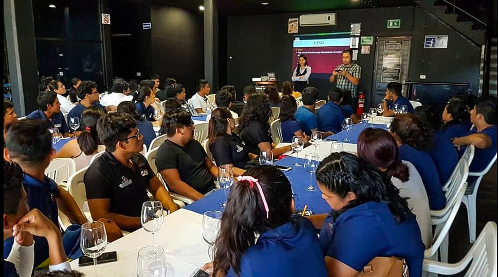 A group of people seated at tables in a dimly lit room listening to presenters near a projection screen.