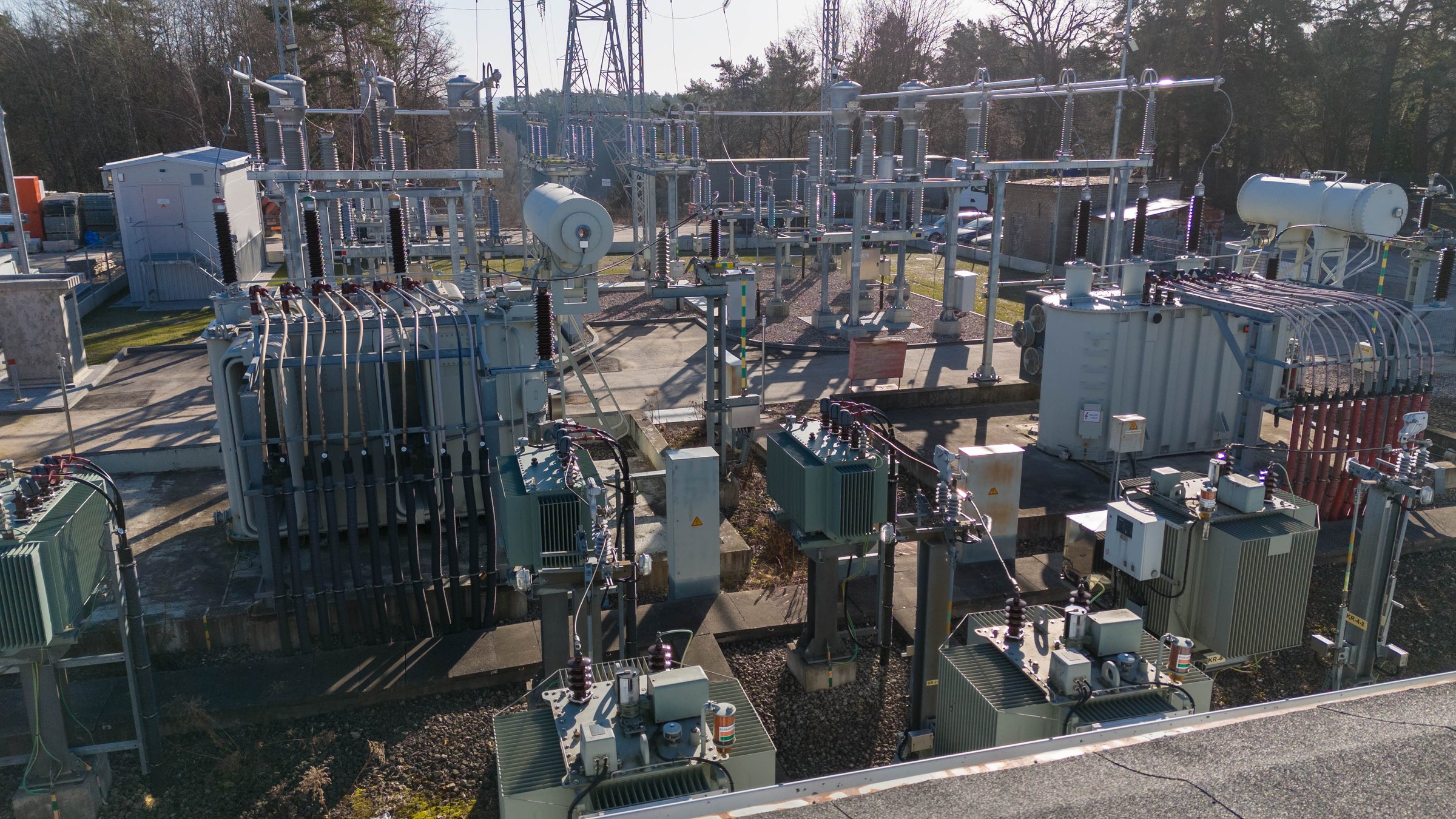 An industrial electrical substation featuring various transformers, circuit breakers, and power lines. The scene is set outdoors with clear skies and sunlight illuminating the equipment.