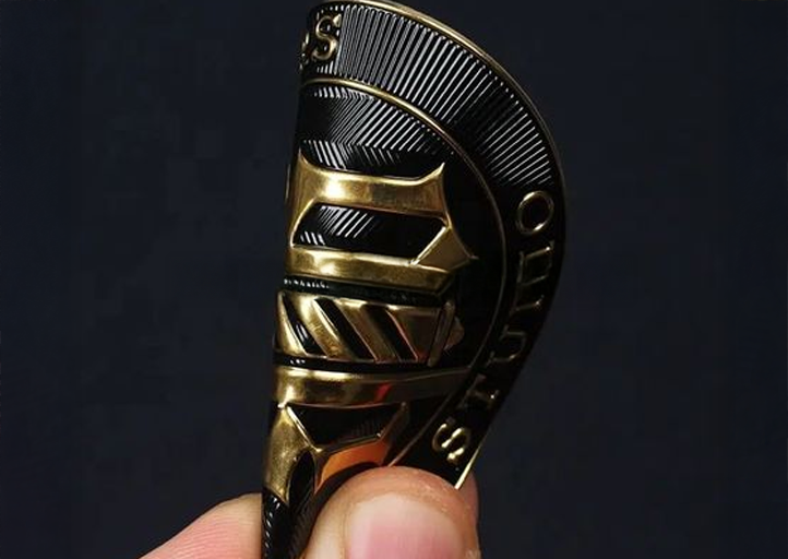 A hand holding a flexible, metallic badge with intricate gold and black detailing against a dark background.