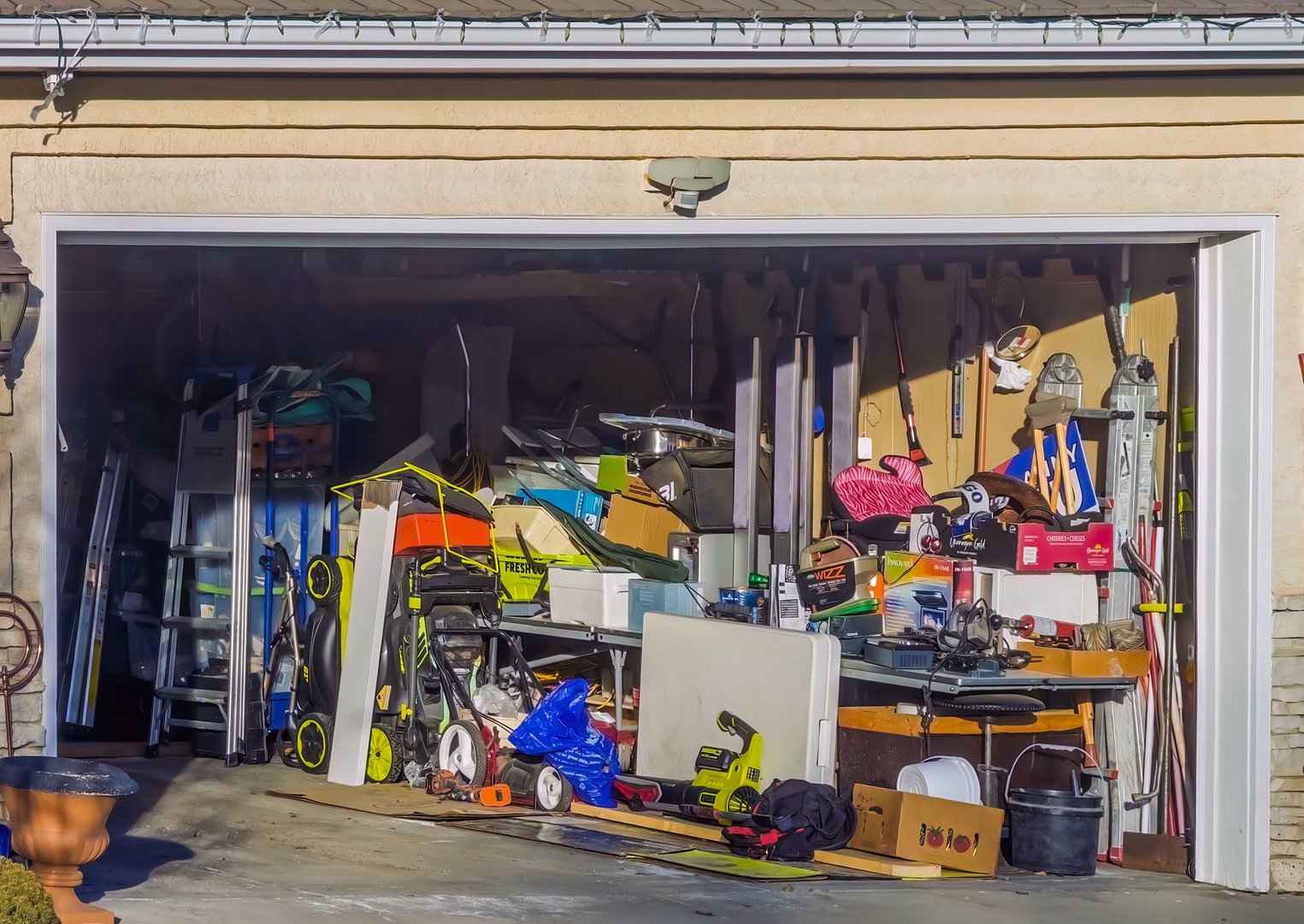 A cluttered suburban garage filled with overflowing household items, lawn equipment, and tools
