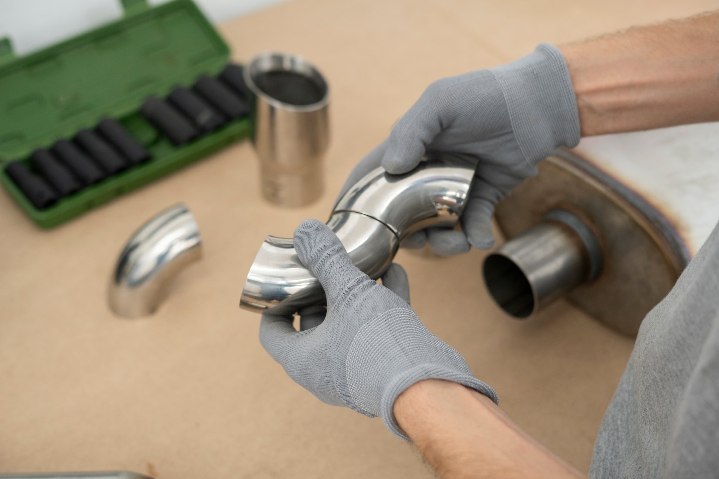 Worker fits together stainless steel elbow pipe joints using gloves in a well-organized workshop. Tools and materials are neatly arranged on a workbench.