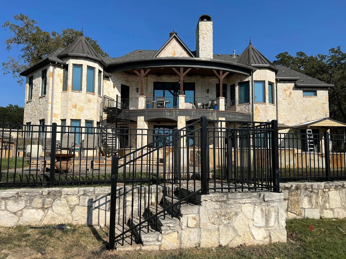 Two-story stone house with turrets, balcony, and iron fence. Set in a grassy area with trees in the background.