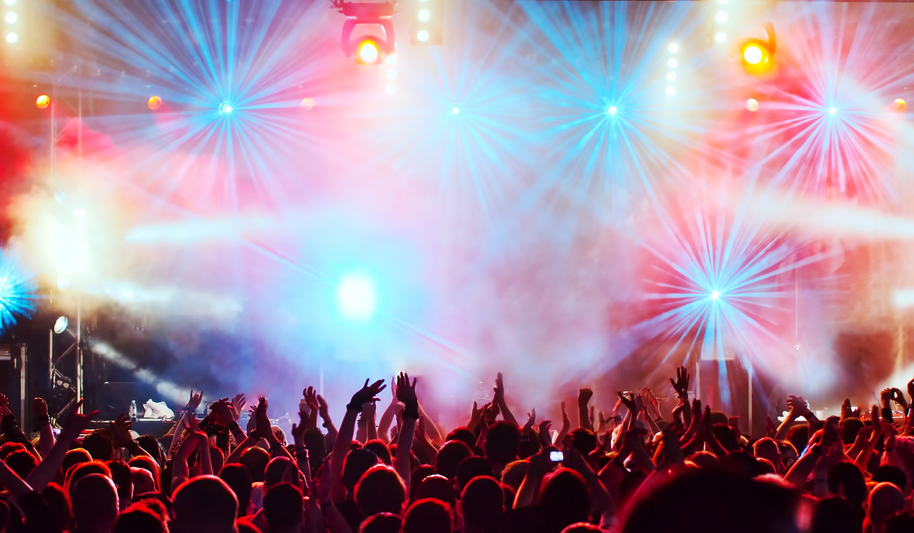 A crowd with raised hands at a concert, illuminated by vibrant red and blue stage lights and fog effects.