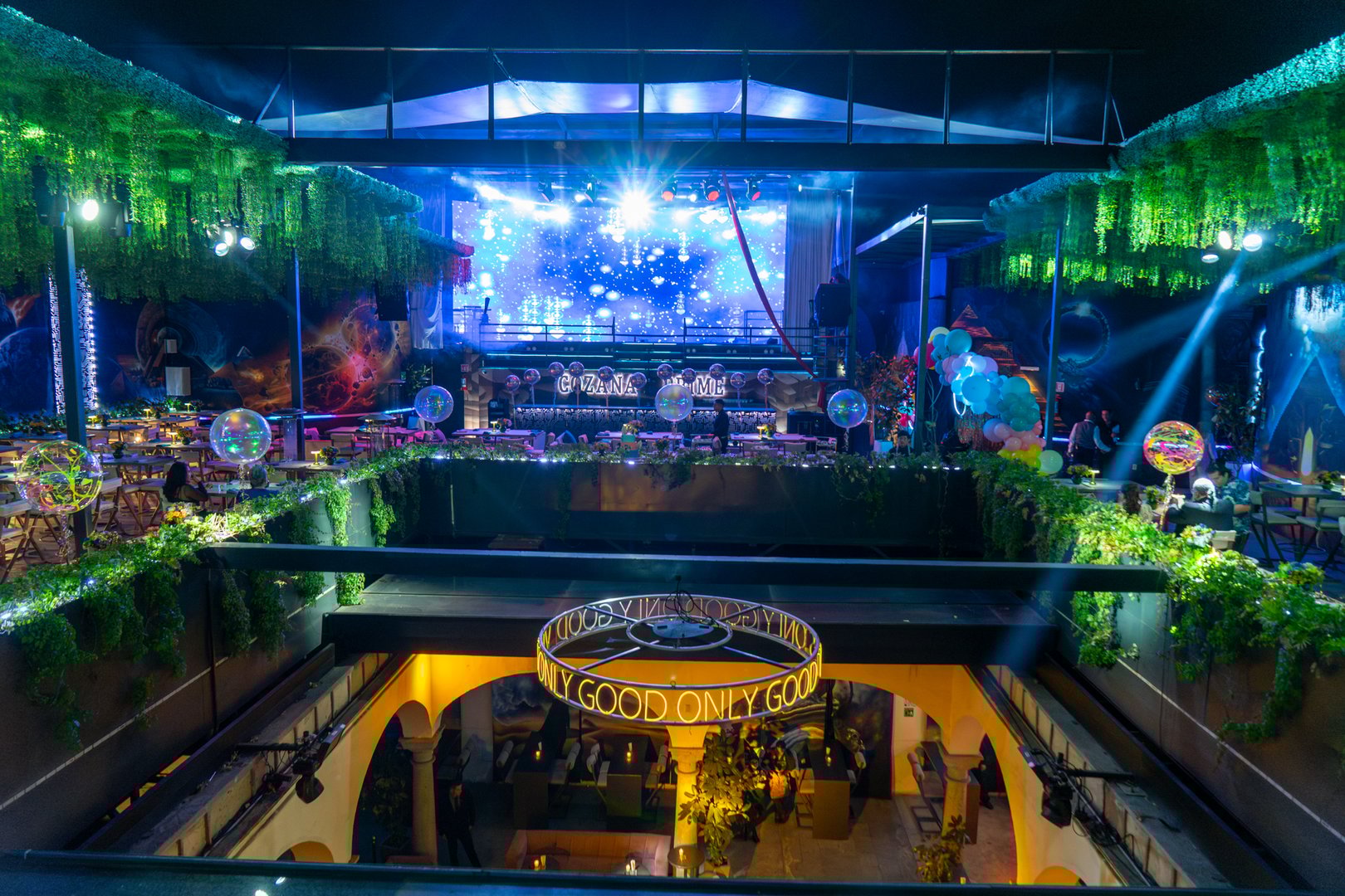 Vibrant rooftop event space with stage, colorful lights, foliage, and tables with illuminated centerpieces at night.