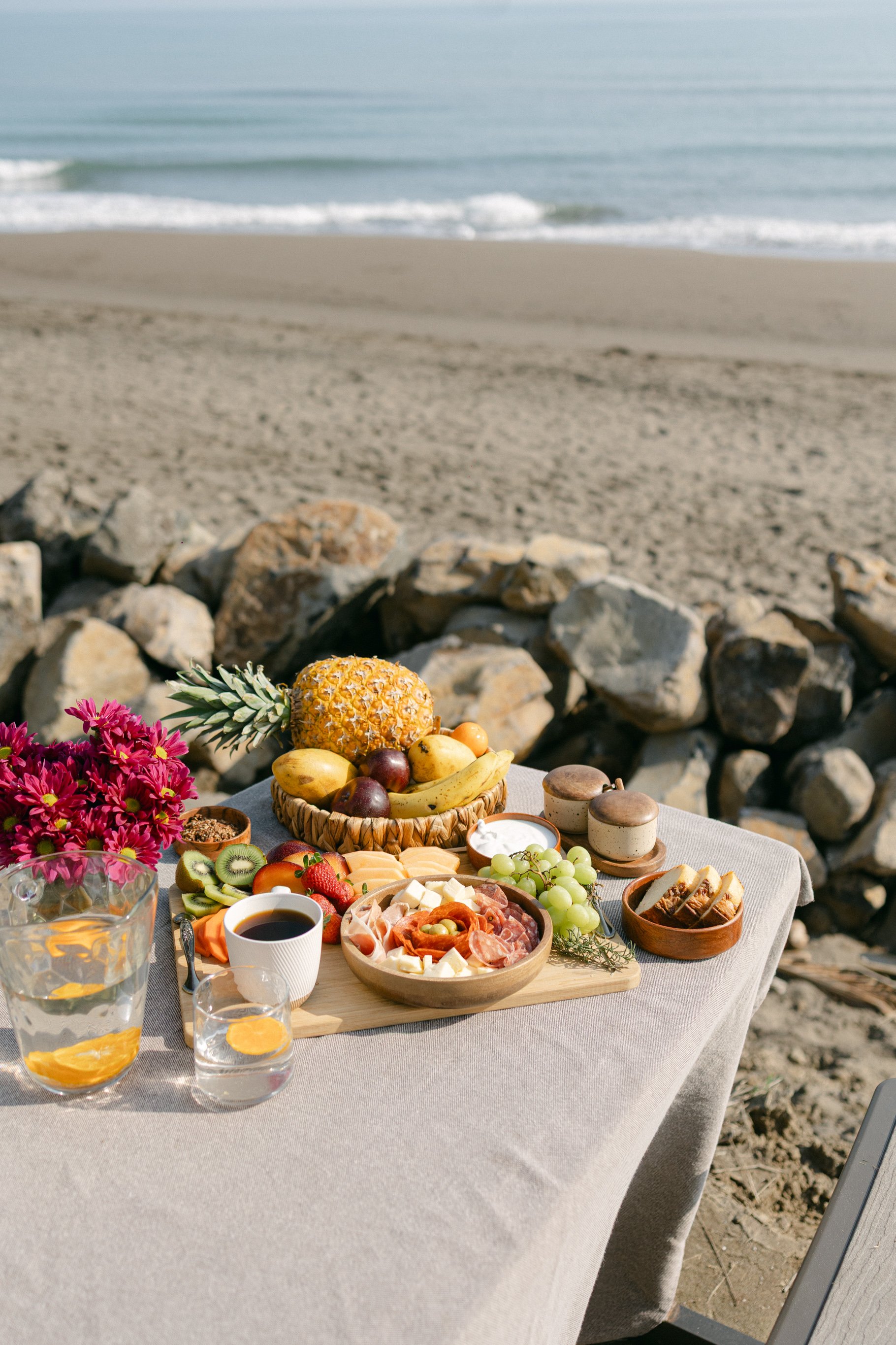 Outdoor picnic by the beach with a spread of fruits, cheese, and flowers on a table, with a view of the ocean.