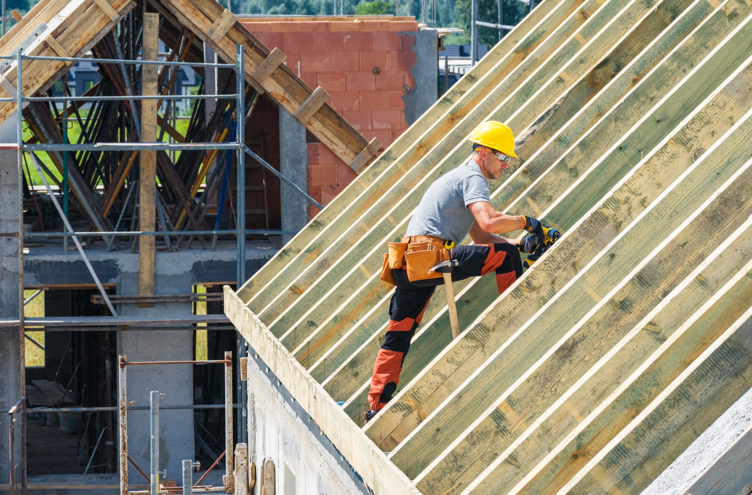 A construction worker is seated on a roof, skillfully securing wooden beams