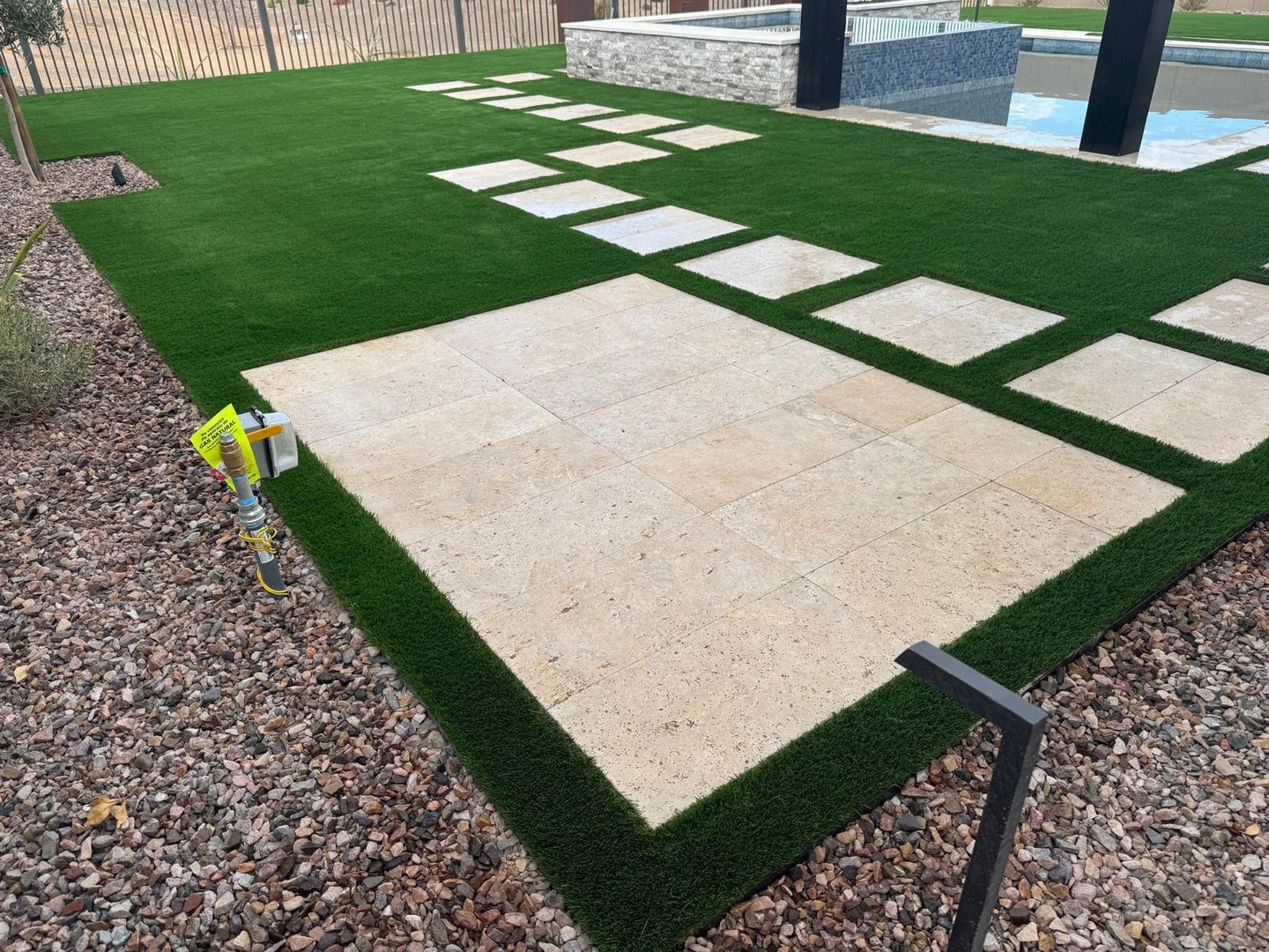 Backyard with artificial grass, stone tiles, gravel border, and a pool in the background.