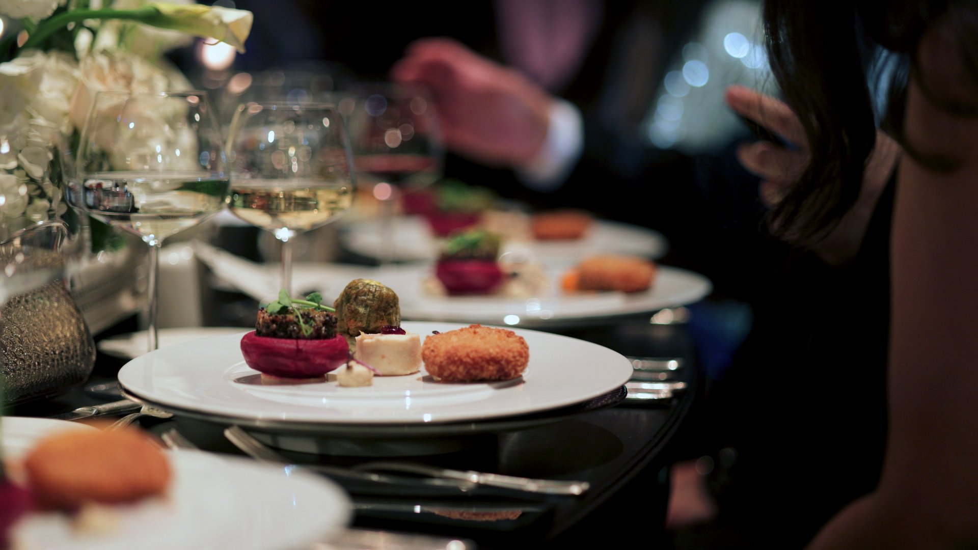 A beautifully plated gourmet dish served at a sophisticated event, accompanied by fine wine and elegant table settings. The dim lighting and luxurious ambiance create an intimate dining atmosphere. Perfect for themes related to fine dining, luxury events, and gastronomy.
Guests indulging in fine wine and exquisite cuisine at an elegantly decorated restaurant. A beautifully plated dinner set on a luxurious table, creating the perfect ambiance for a memorable wedding celebration.