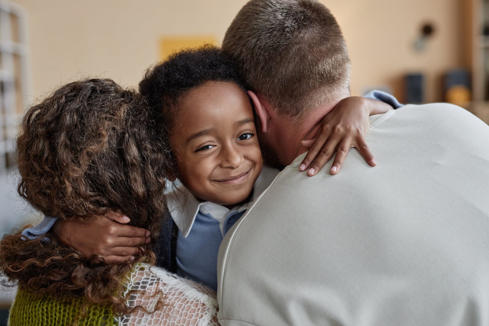 Portrait shot of little happy Black boy giving tender embrace to mother and father while supporting parents caring for adopted kid in childs bedroom