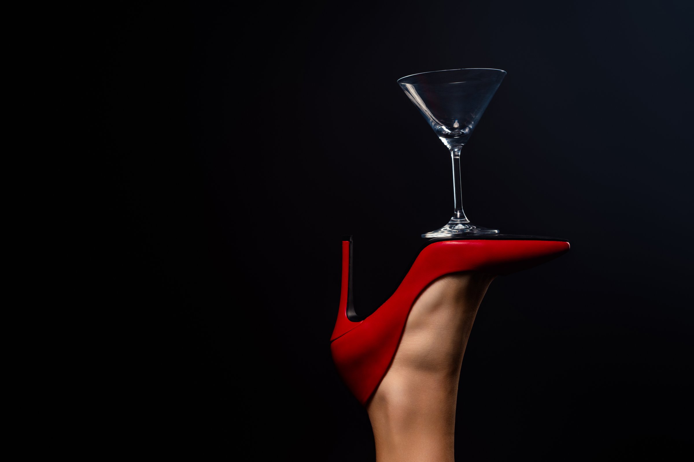 A close-up artistic shot of a red high heel shoe balancing a martini glass, emphasizing elegance, glamour, and sophistication, set against a black backdrop for striking contrast and mystique.