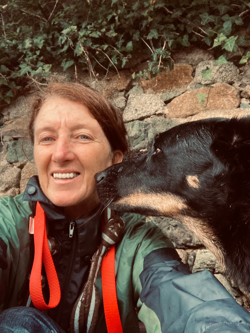 Person in a raincoat smiling with a black and tan dog licking their face, in front of stone and ivy-covered wall.