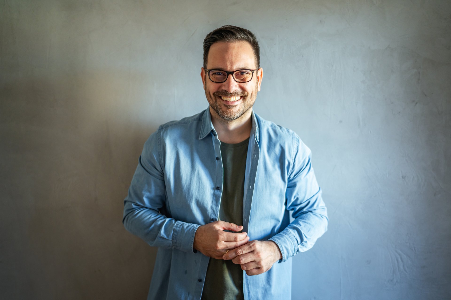 Man with a happy expression and casual attire wearing eyeglasses while standing confidently against a plain gray background, portraying a cheerful and approachable personality