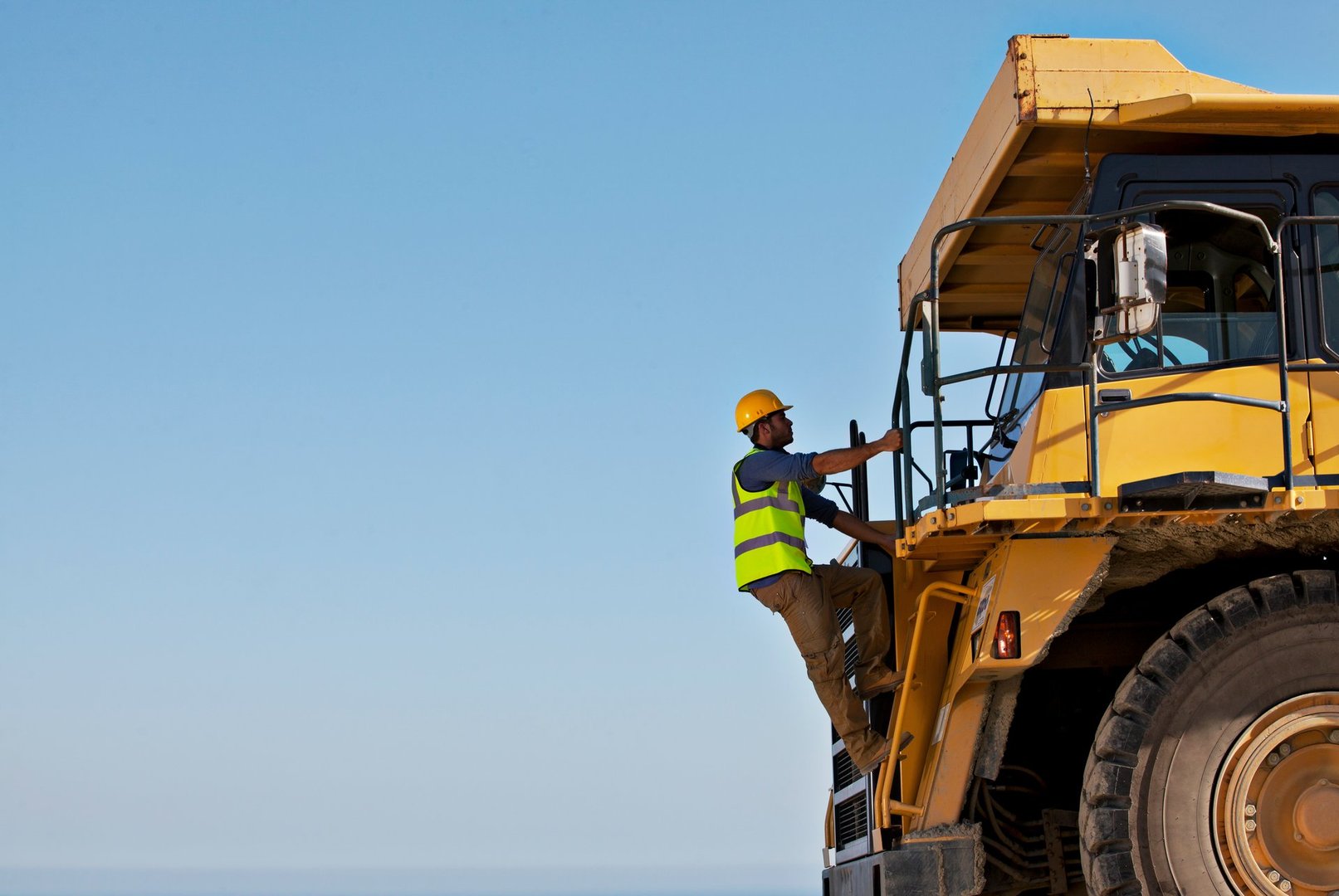 Worker in safety gear climbs large yellow construction vehicle against a clear blue sky.