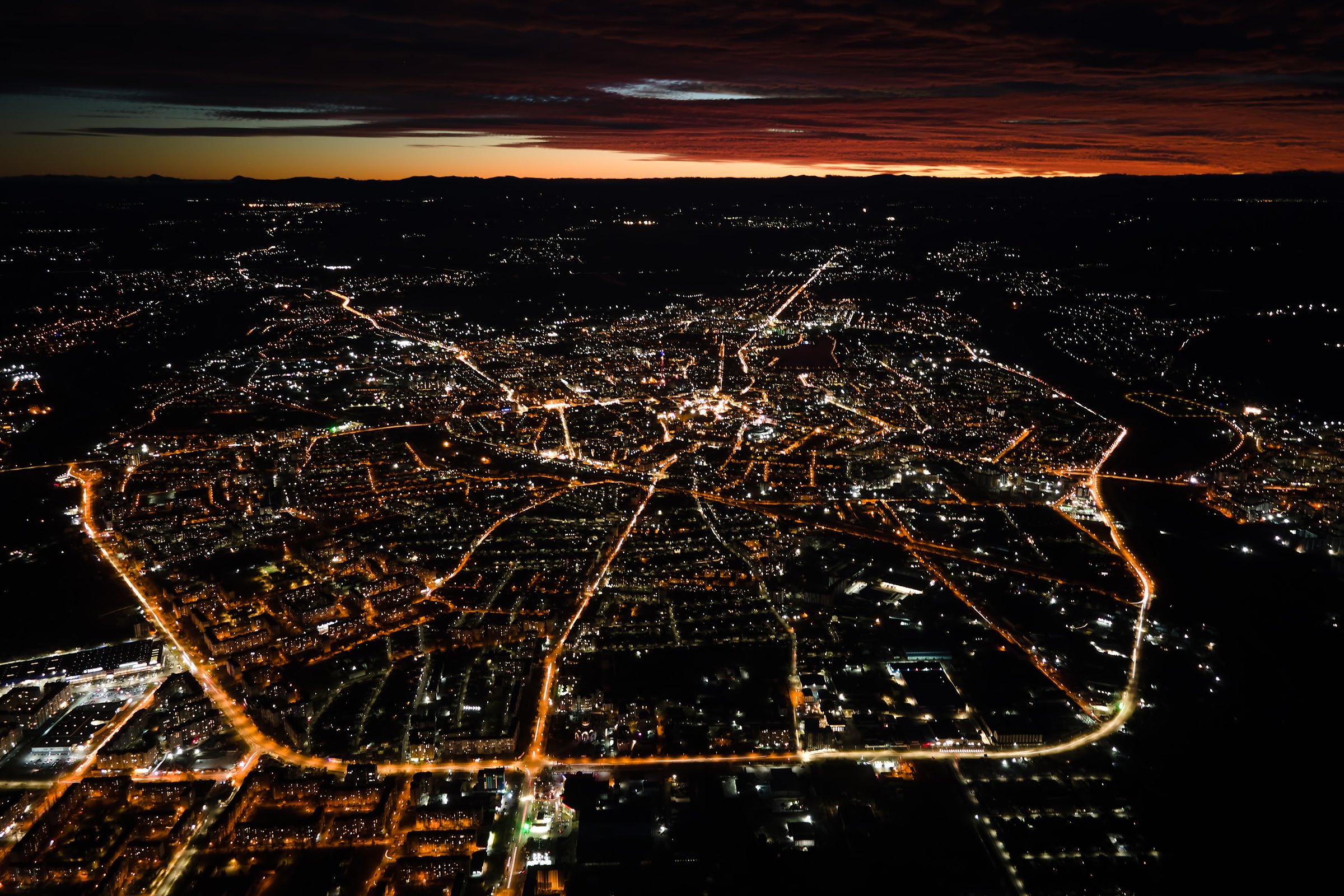 Aerial view from airplane window of buildings and bright illuminated streets in city residential area at night. Dark urban landscape at high altitude.
