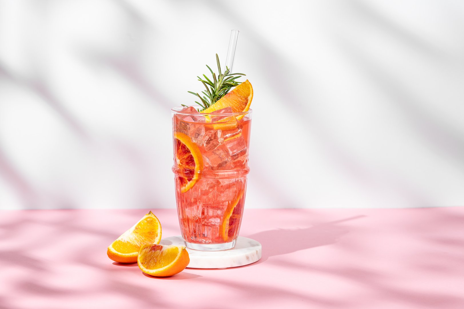 Blood orange cocktail with ice and rosemary on a marble stand on a pink white background with shadows. Summer citrus drink concept for cafe menu advertising banner. Front view, copy space