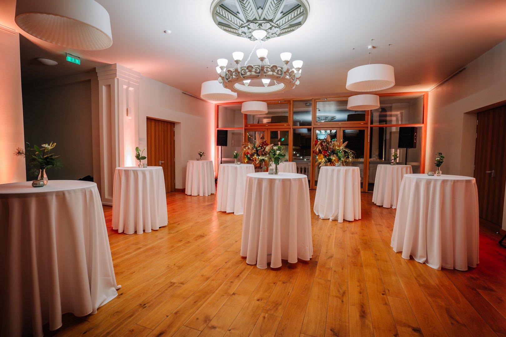 A modern, elegant event room with round cocktail tables covered in white cloth, decorated with floral centerpieces and lit warmly.