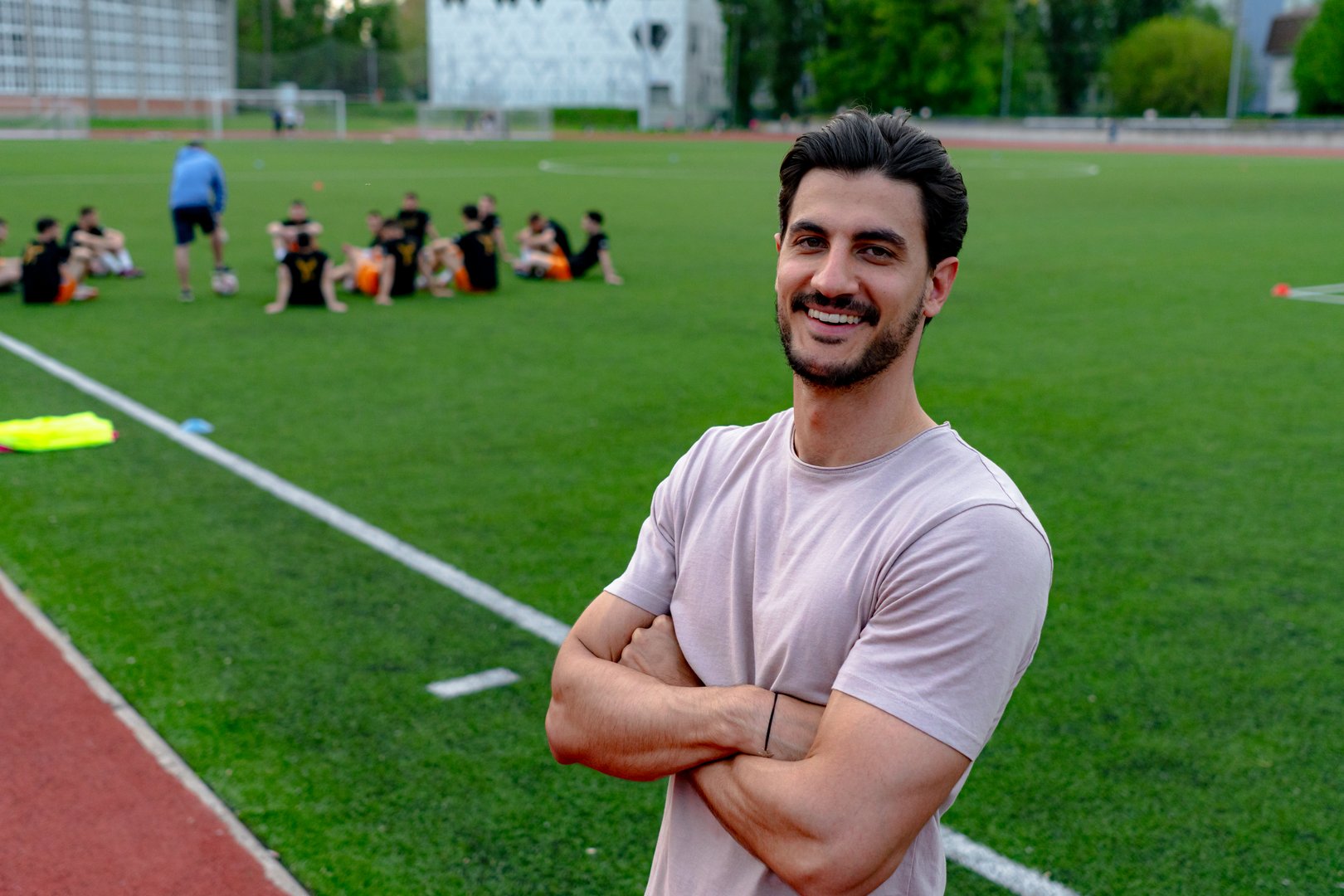 Portrait of a young smiling soccer coach standing with arms crossed on a playing field while his team is training