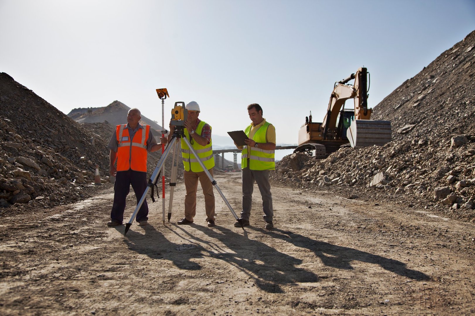 Three surveyors in safety vests use equipment at a construction site with an excavator in the background.