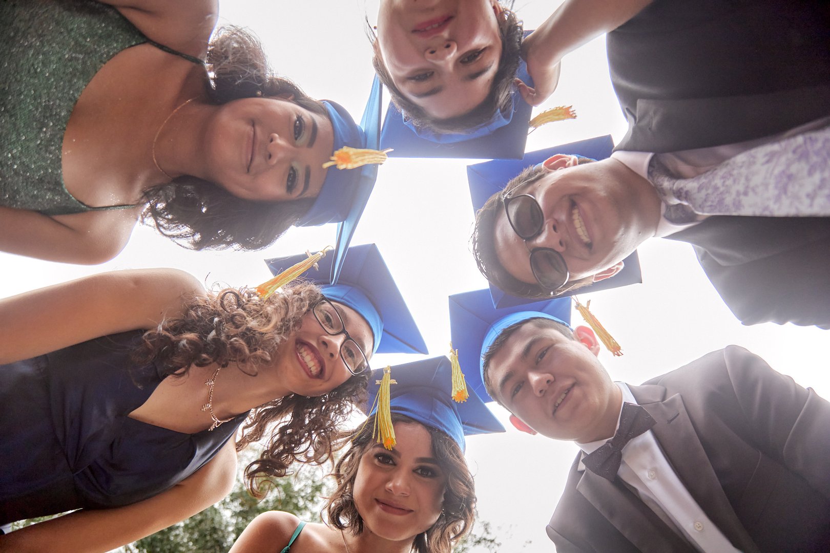 Group of graduates in blue caps and gowns huddled together, looking down at the camera with smiles.