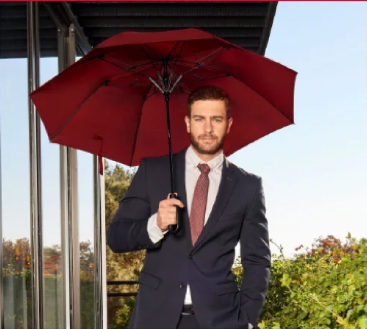 Man in a light shirt carrying a blue bag with a strap, walking near a glass door with a view of trees in the background.