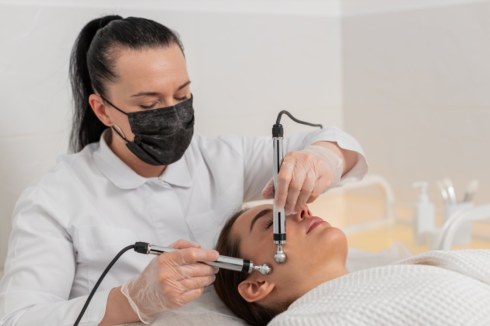 Caucasian girl receives targeted microcurrent therapy on temple, jawline and cheek during rejuvenation session. Young woman lies calm while cosmetologist uses tools to improve skin tone and firmness