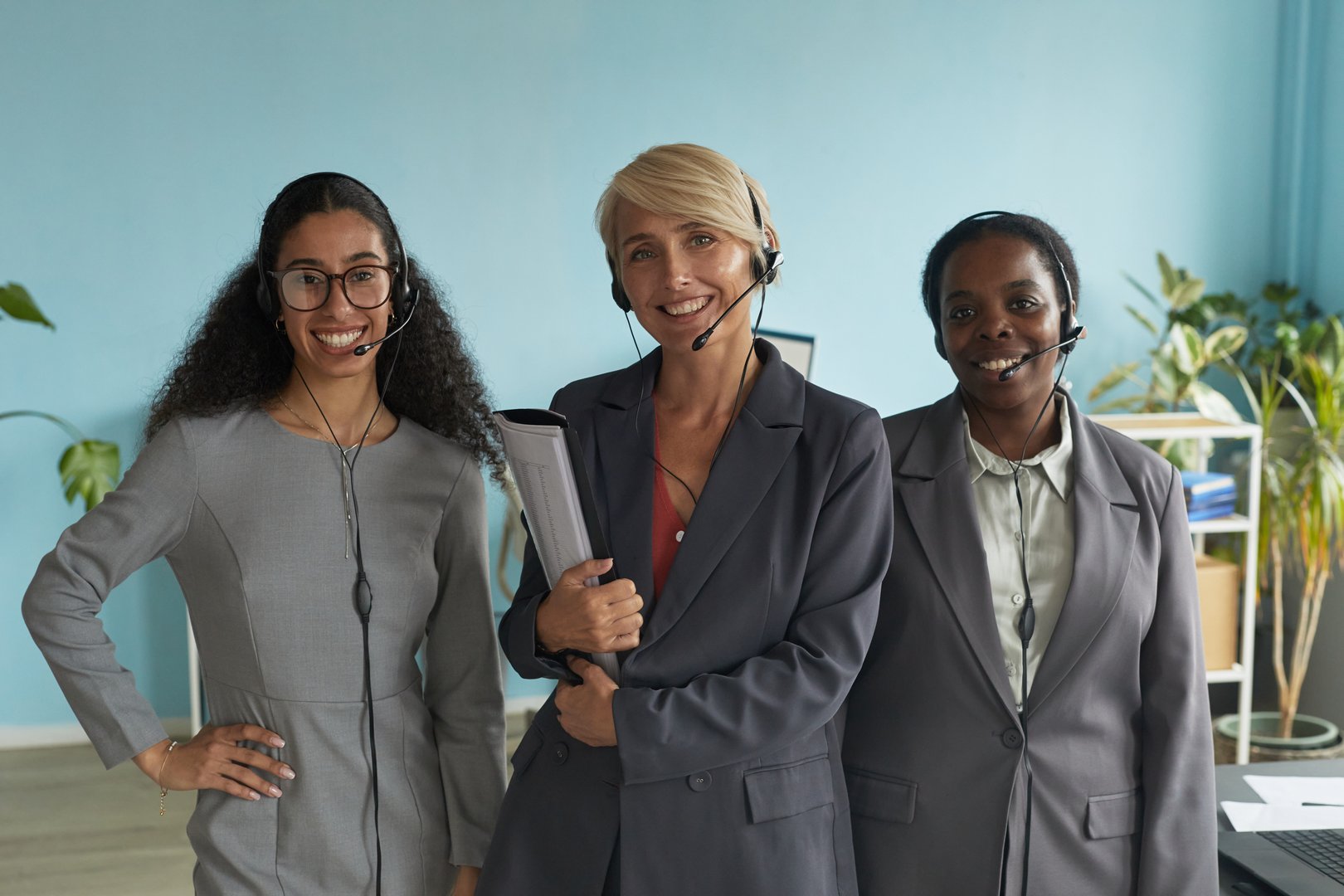 Three diverse women standing together wearing headsets, smiling at camera, demonstrating teamwork in modern office environment