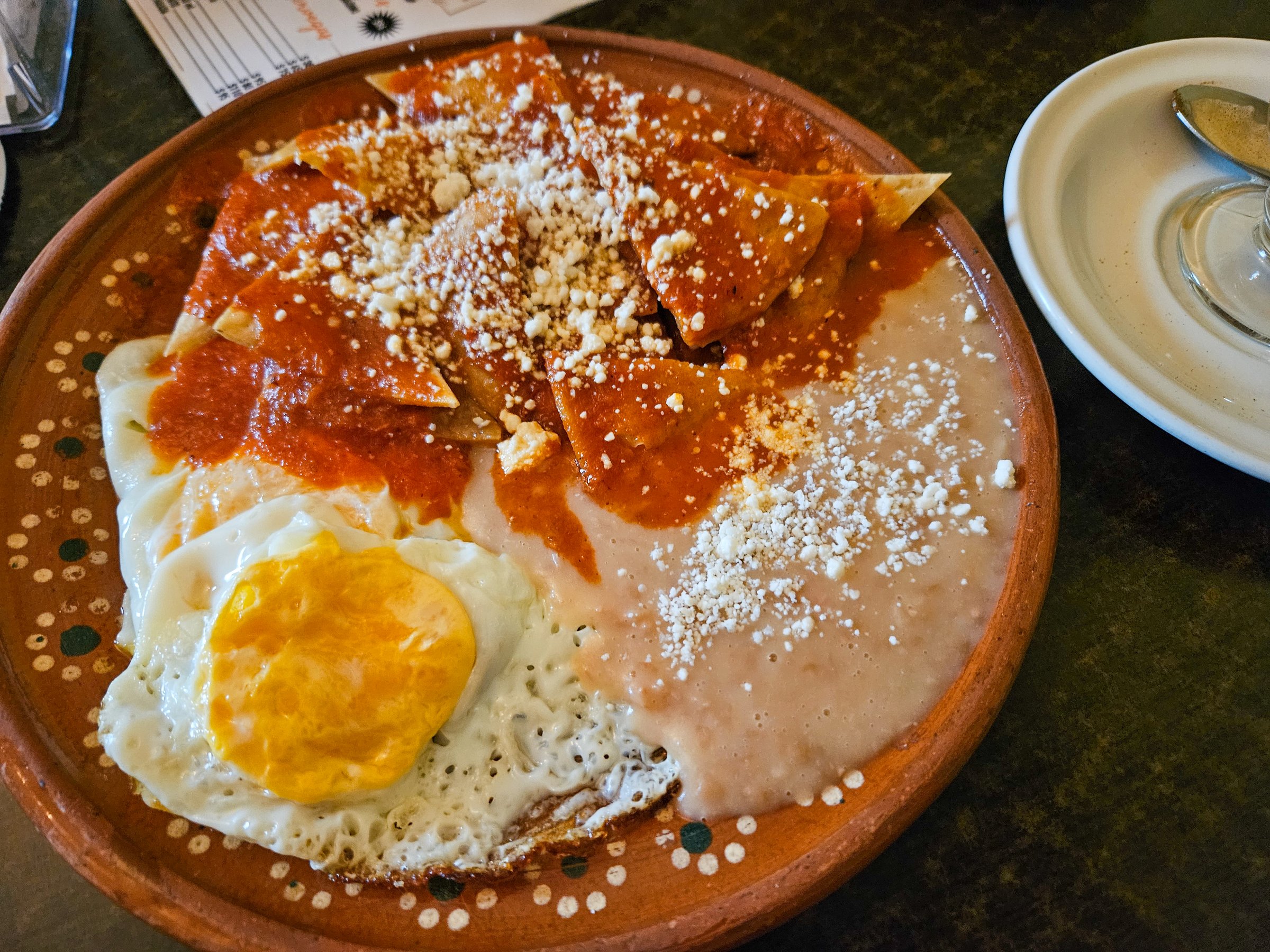 A delicious traditional Mexican breakfast of chilaquiles with a sunny-side-up fried egg and refried beans on a rustic clay plate. Topped with red sauce and crumbled cheese