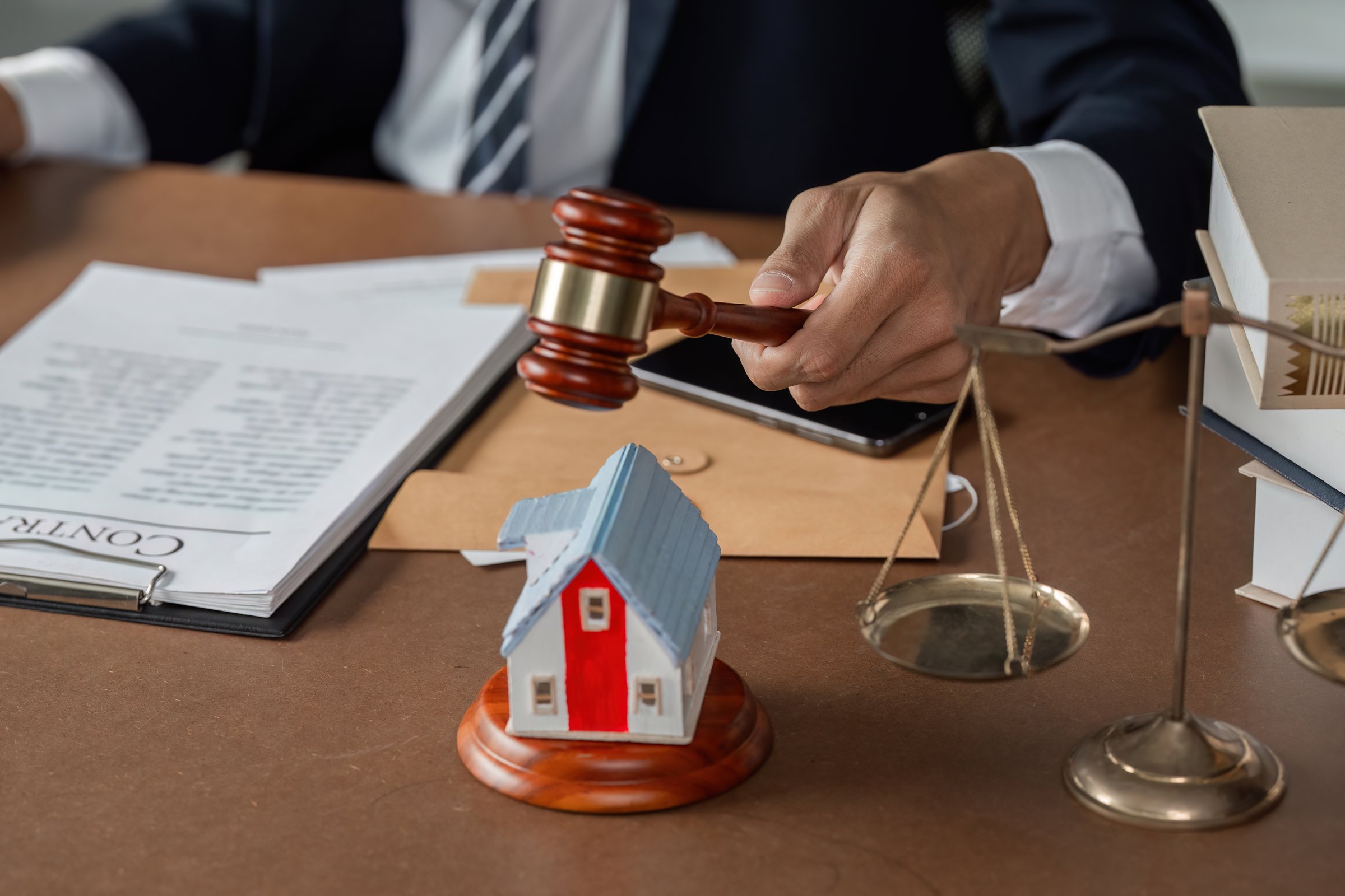 A lawyer is striking a gavel on the desk next to a model house and scales of justice, while reviewing legal documents in a modern office.