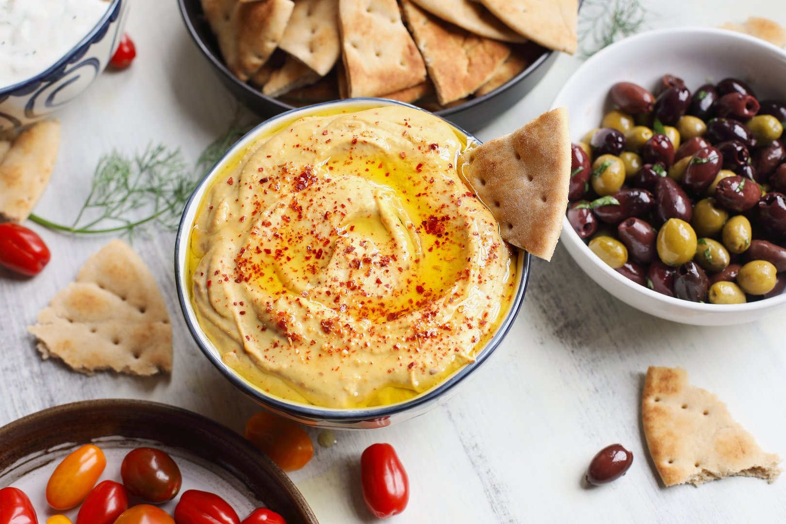 Overhead view of mezze platter of hummus and pita bread surrounded by fresh tomatoes, olives, and vegan tzatziki dip over a white rustic table.