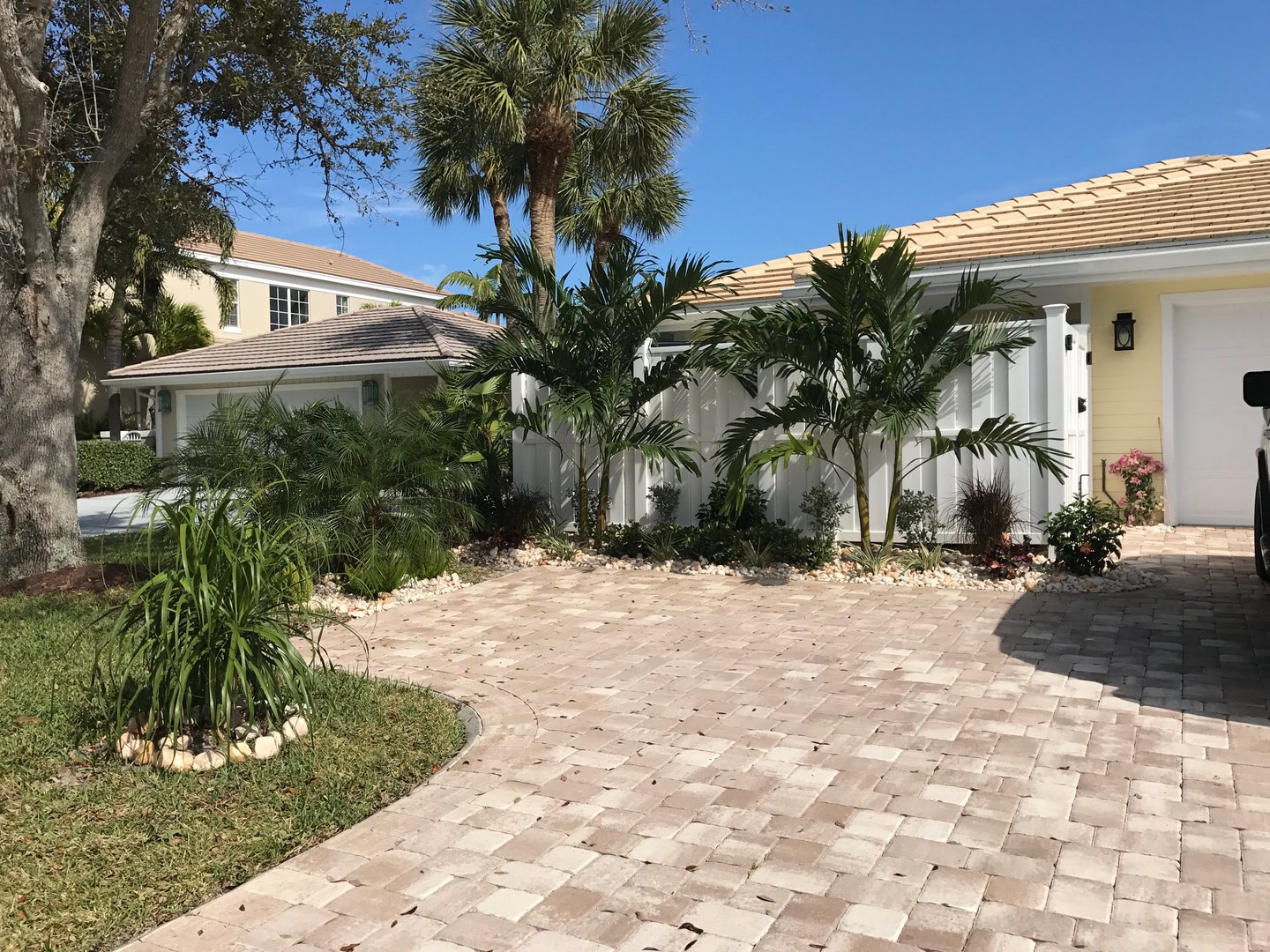Brick driveway leading to a white garage, surrounded by palm trees and a well-maintained garden under a clear blue sky.