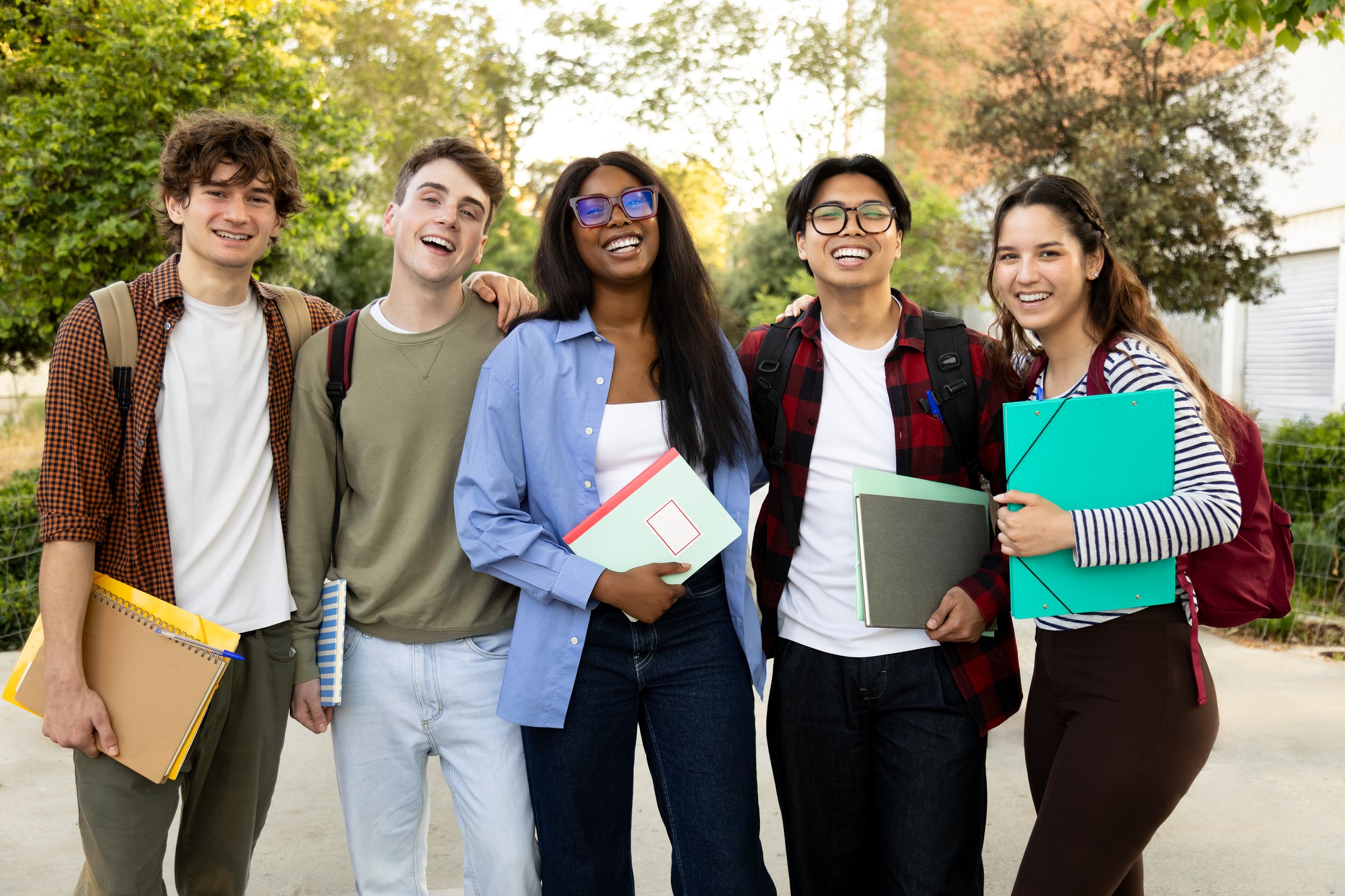 Group of cheerful multi-ethnic students standing together on campus, holding notebooks and smiling at camera