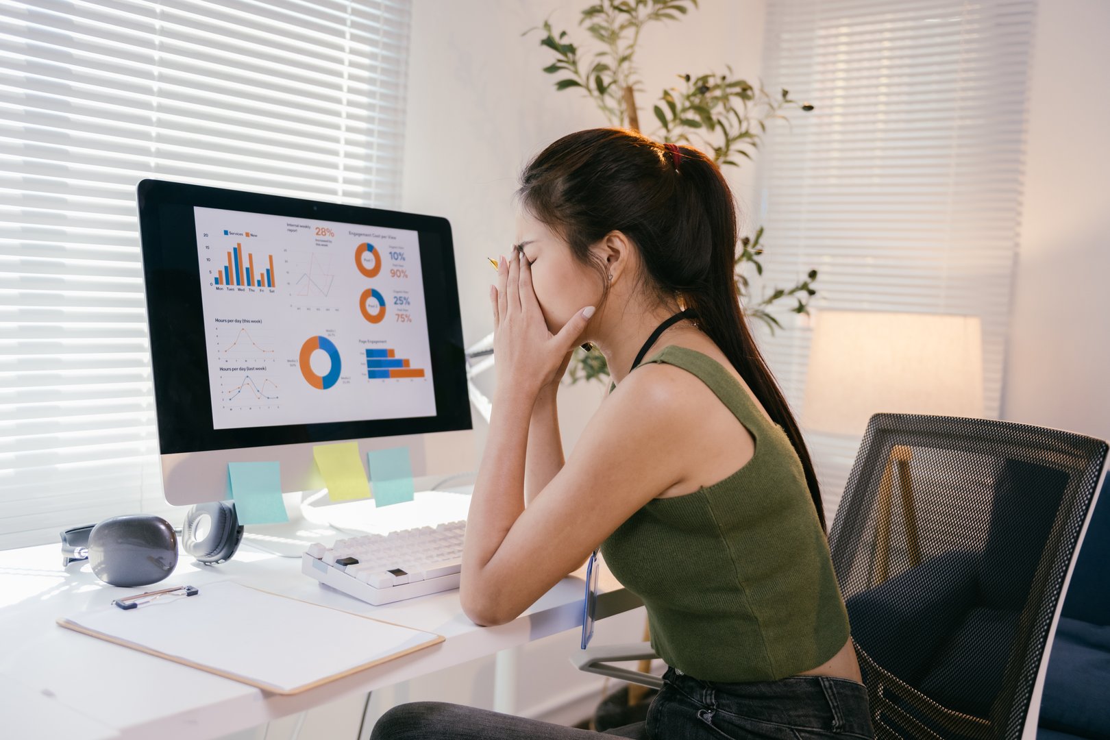 Young businesswoman is sitting at her desk, covering her face with her hands, feeling stressed and overwhelmed by the disappointing charts displayed on her computer screen