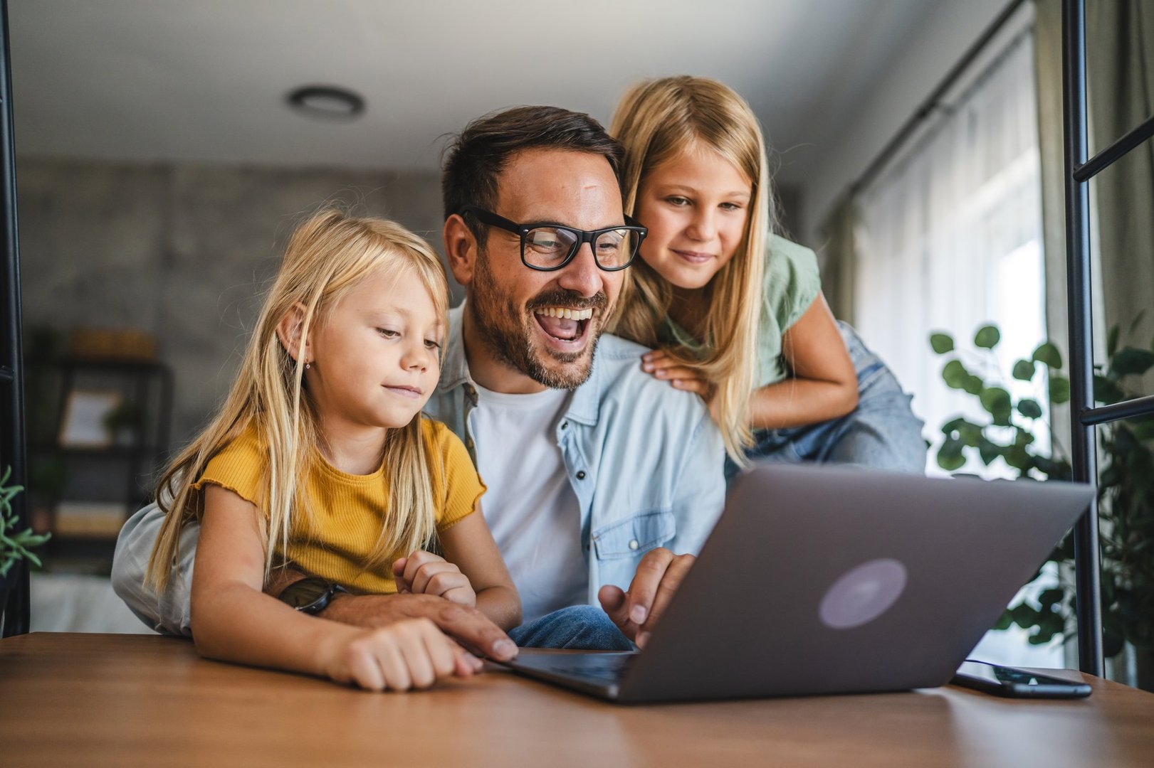 A joyful father with glasses and his two daughters smile while engaging with a laptop at a wooden table in a cozy home, creating bonding and learning memories.