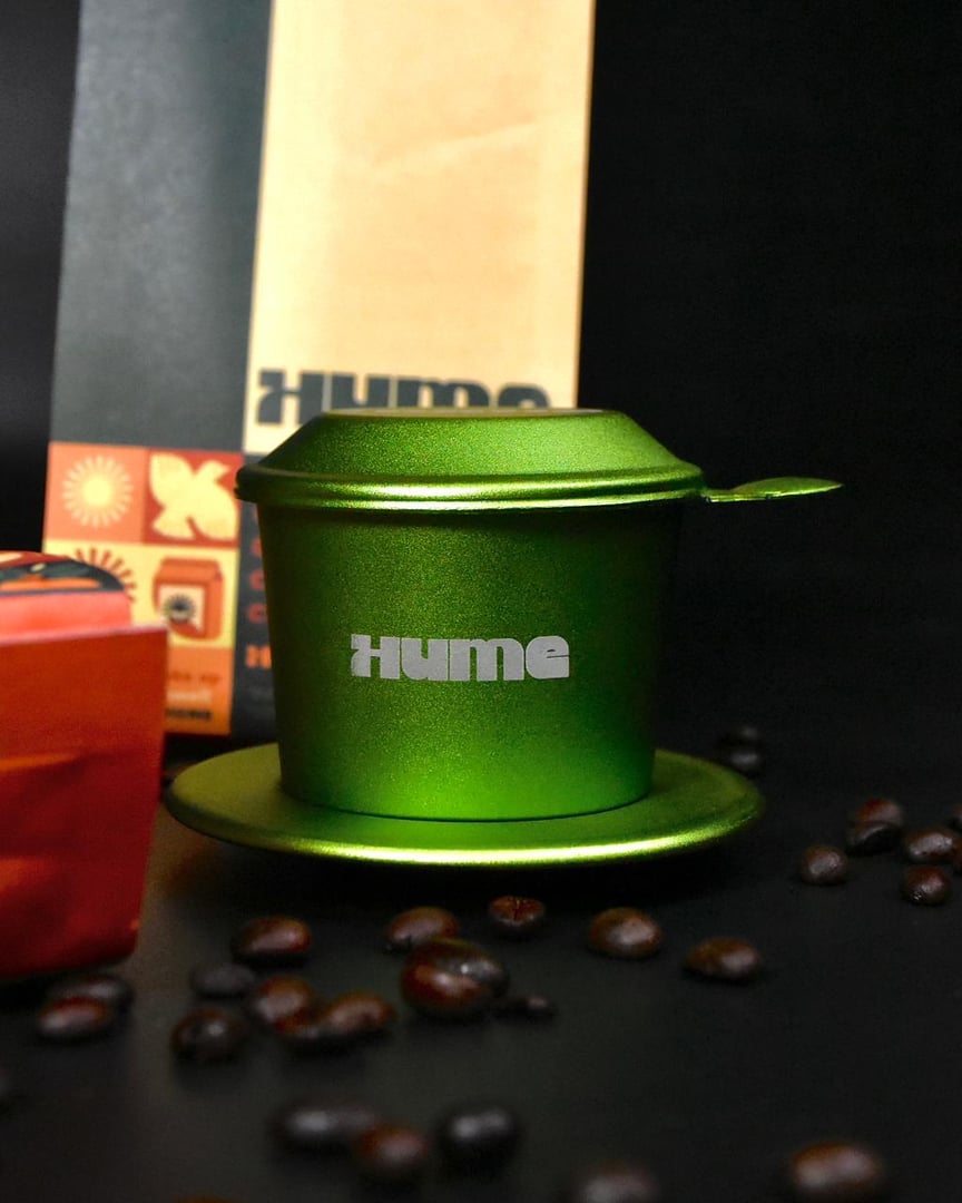 Green metal coffee dripper on a saucer with "Hume" printed, surrounded by coffee beans, with packaging in the background.