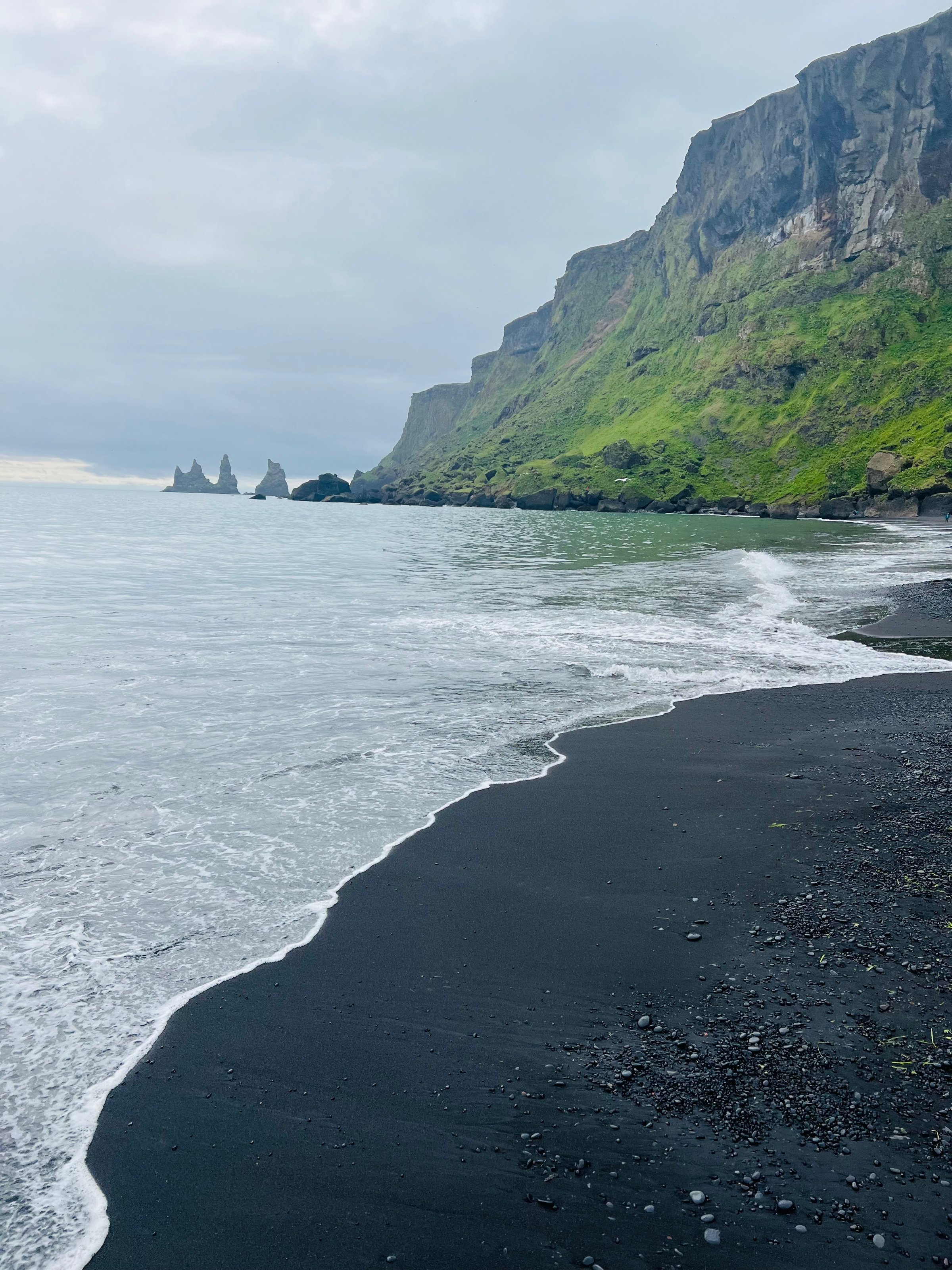 Black sand beach with wave, rocky sea stack formations, and steep green cliffs under a cloudy sky.