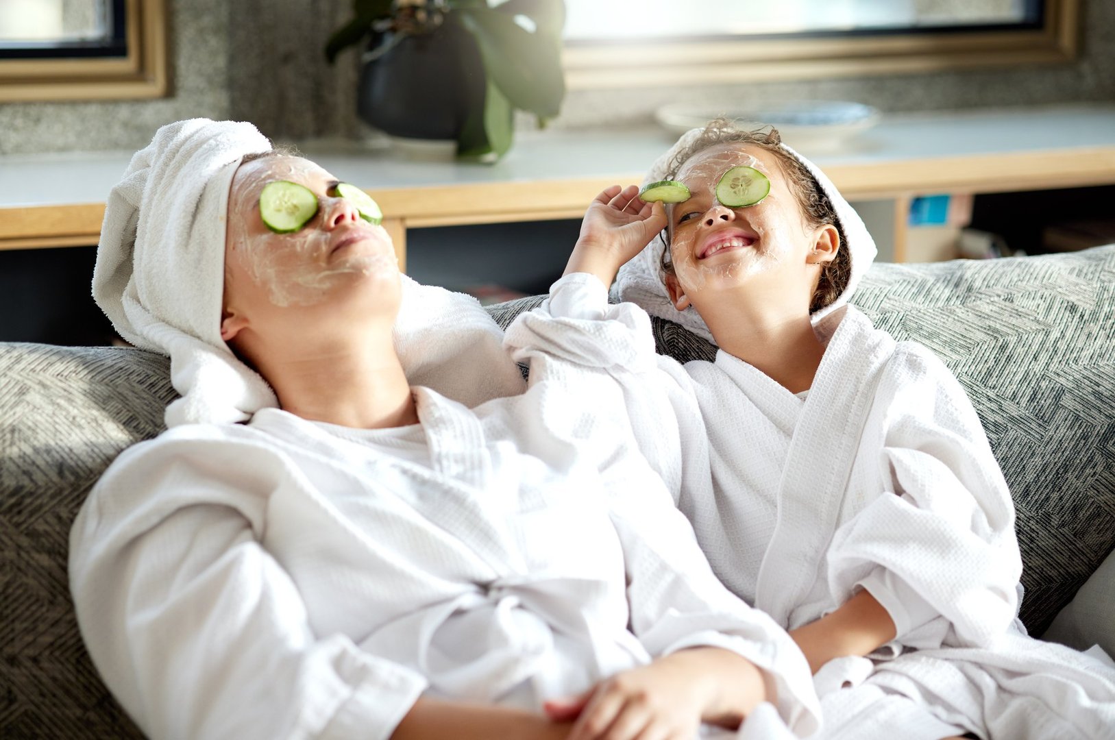 Fresh skincare, face mask and healthy skin treatment for mother and daughter home spa day. Fun, smiling and playful child and single parent relaxing with cucumber over their eyes in grooming routine
