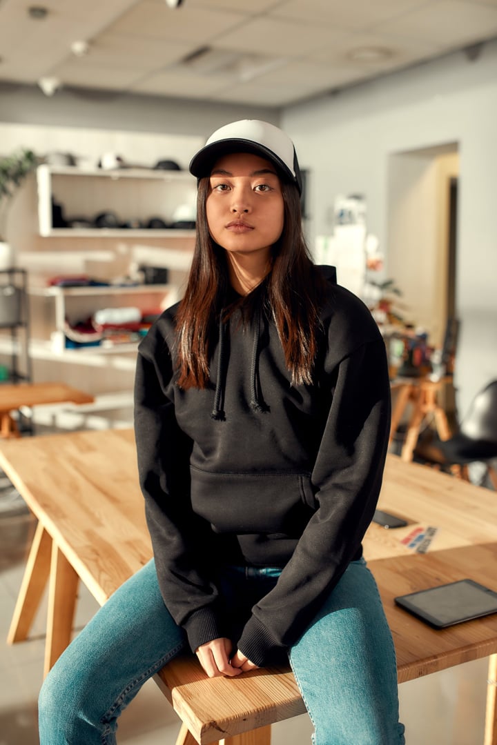 Cheerful female worker looking at camera while posing in custom apparel, baseball cap and hoodie. Young woman working at custom T-shirt, clothing printing company. Vertical shot