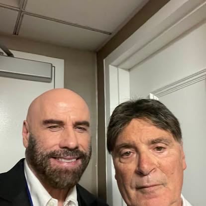 Two men posing together indoors, one with a beard and the other with short dark hair, standing in front of a white door.