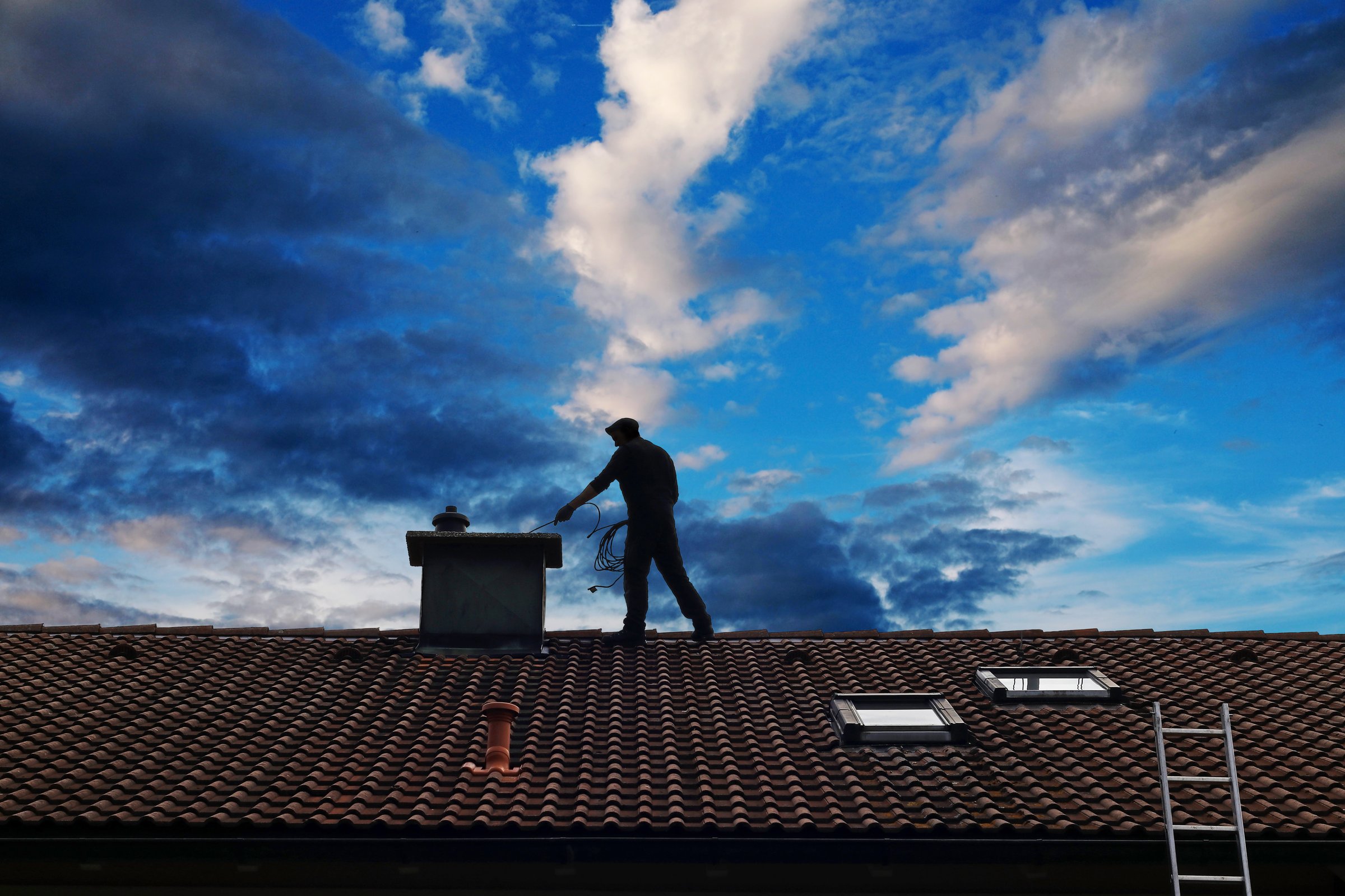 A chimney sweep cleans the chimney on the roof of a house in front of a beautiful sky