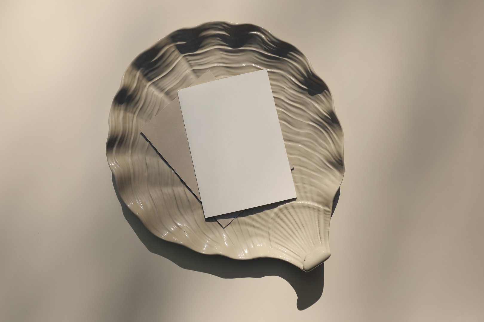 Modern summer stationery still life. Blank greeting card, invitation mock up scene with oyster shell ceramic plate. Beige table background in sunlight, long shadows, flat lay, top view. Vacation, ocean concept.