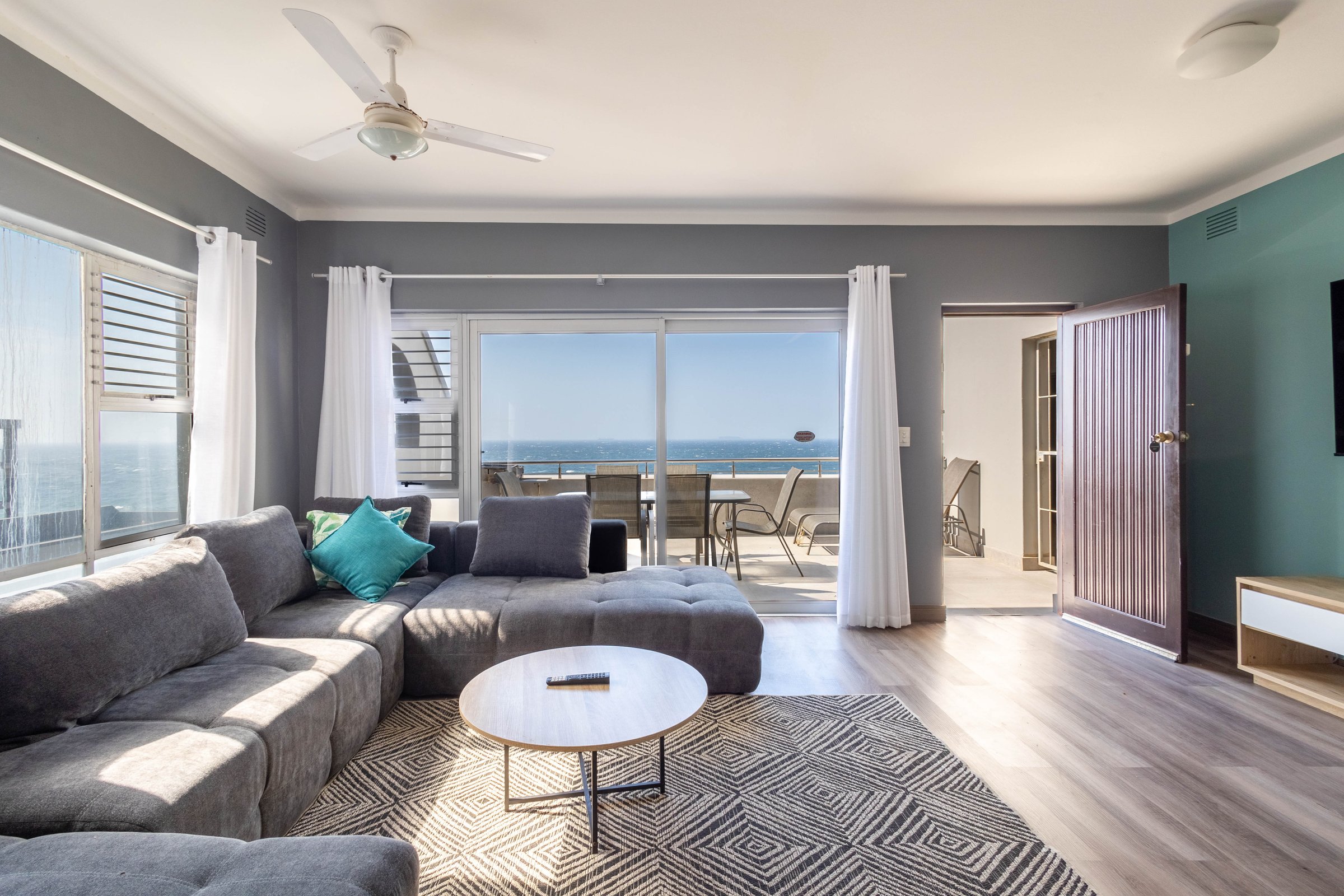Modern living room with a gray sectional sofa, ceiling fan, open door, balcony view of the ocean, and wooden flooring.
