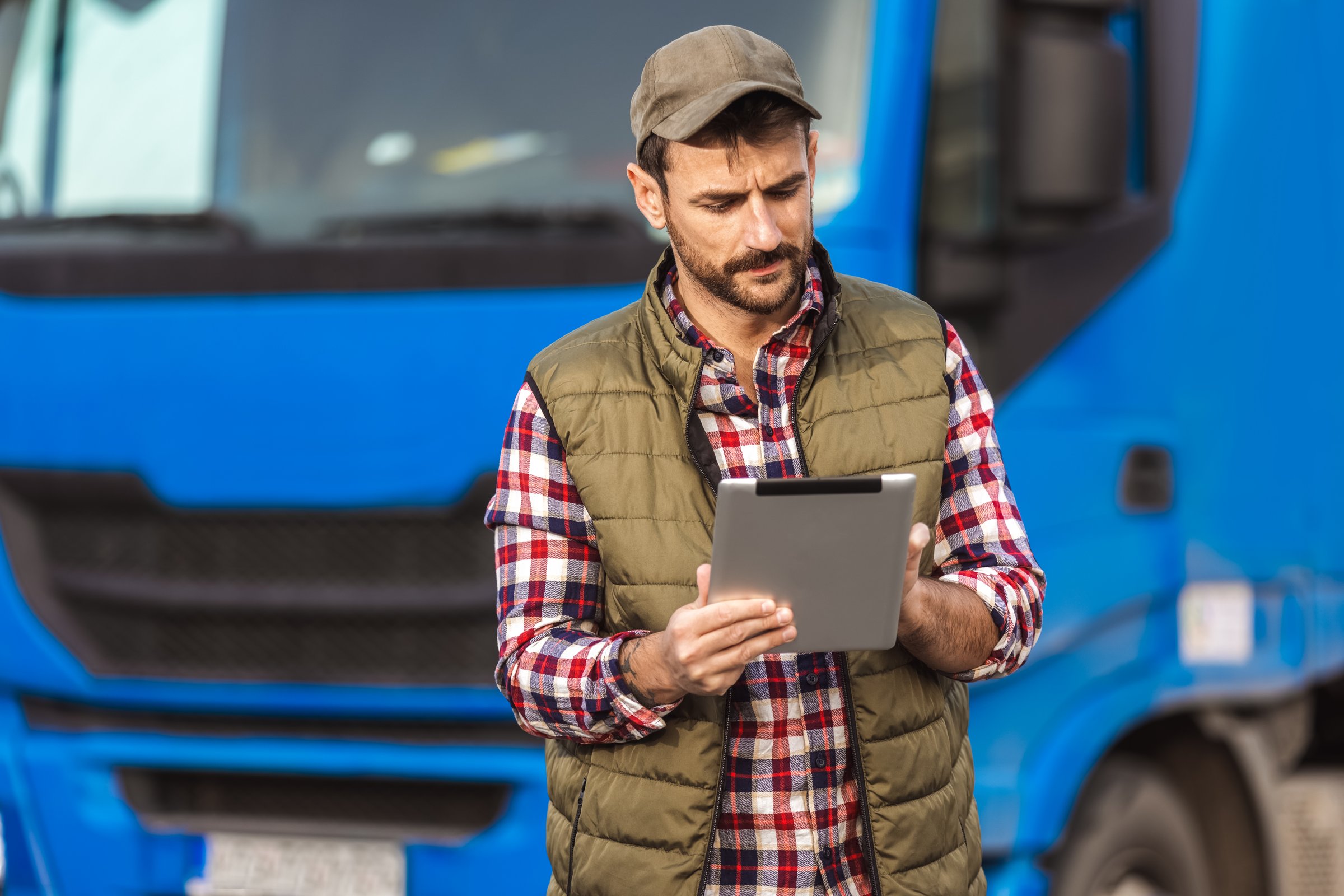 Happy confident male driver standing in front on his truck, using his tablet.