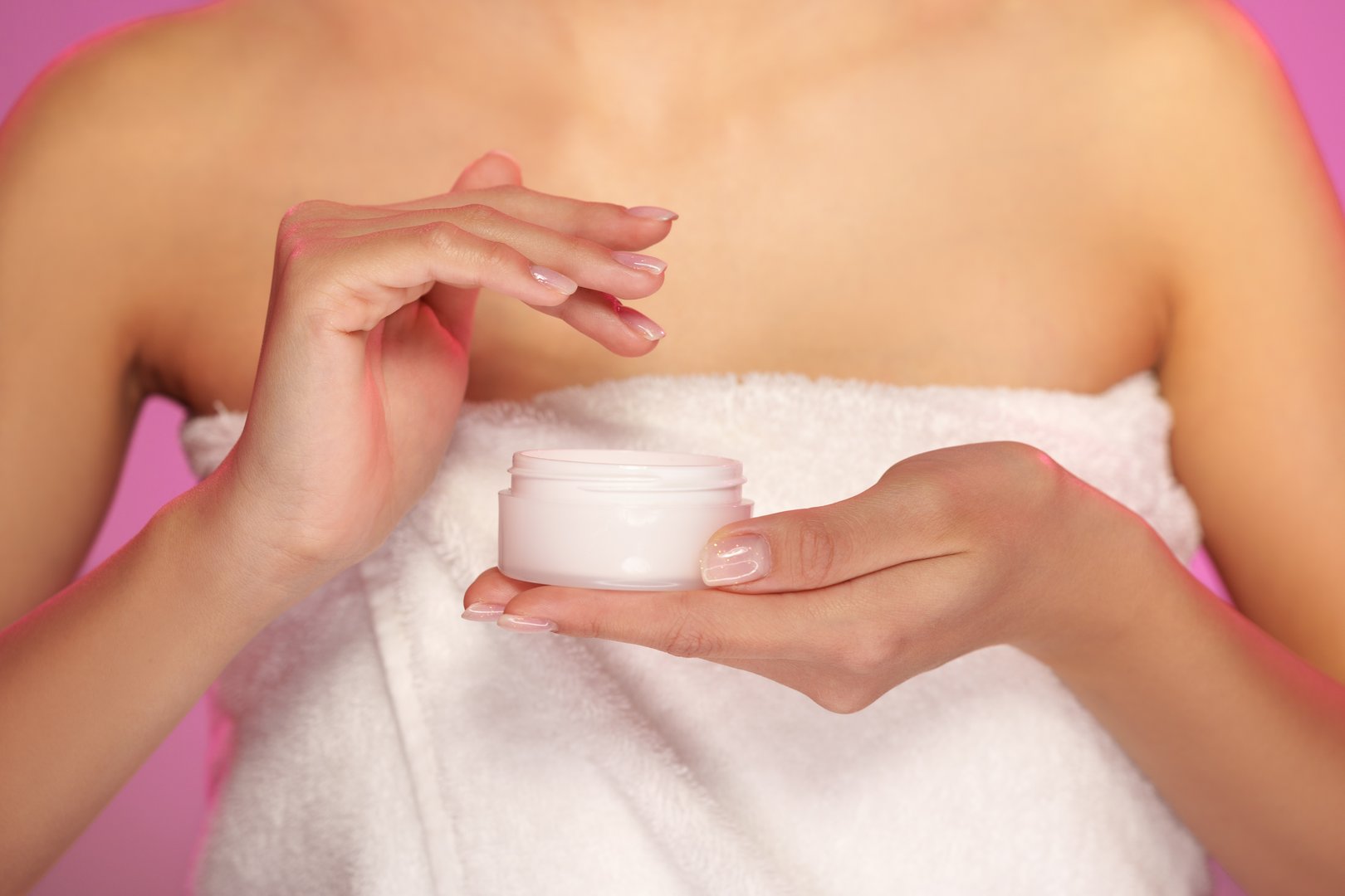 A person wrapped in a towel is applying cream from a small jar, emphasizing skincare practices in a bright, minimalistic environment.