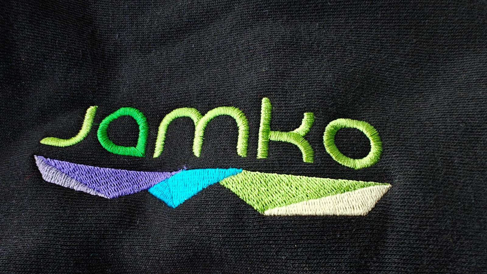 Colorful embroidery of the word JAMKO with abstract geometric shapes on a dark fabric background.